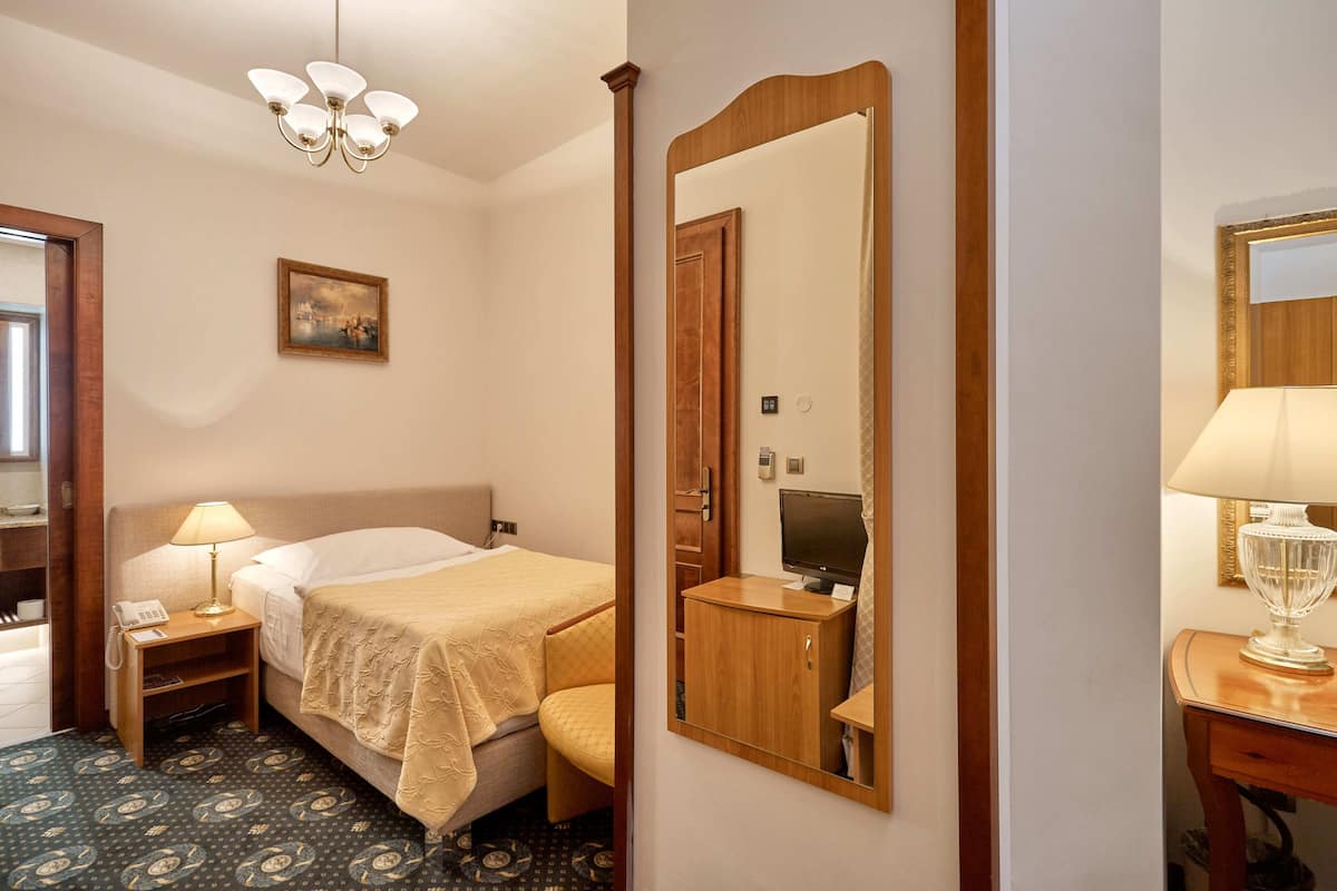 Ein Hotelzimmer mit Bett, Nachttisch mit Lampe, Spiegel, einem Wandbild und einem gemusterten Teppich.