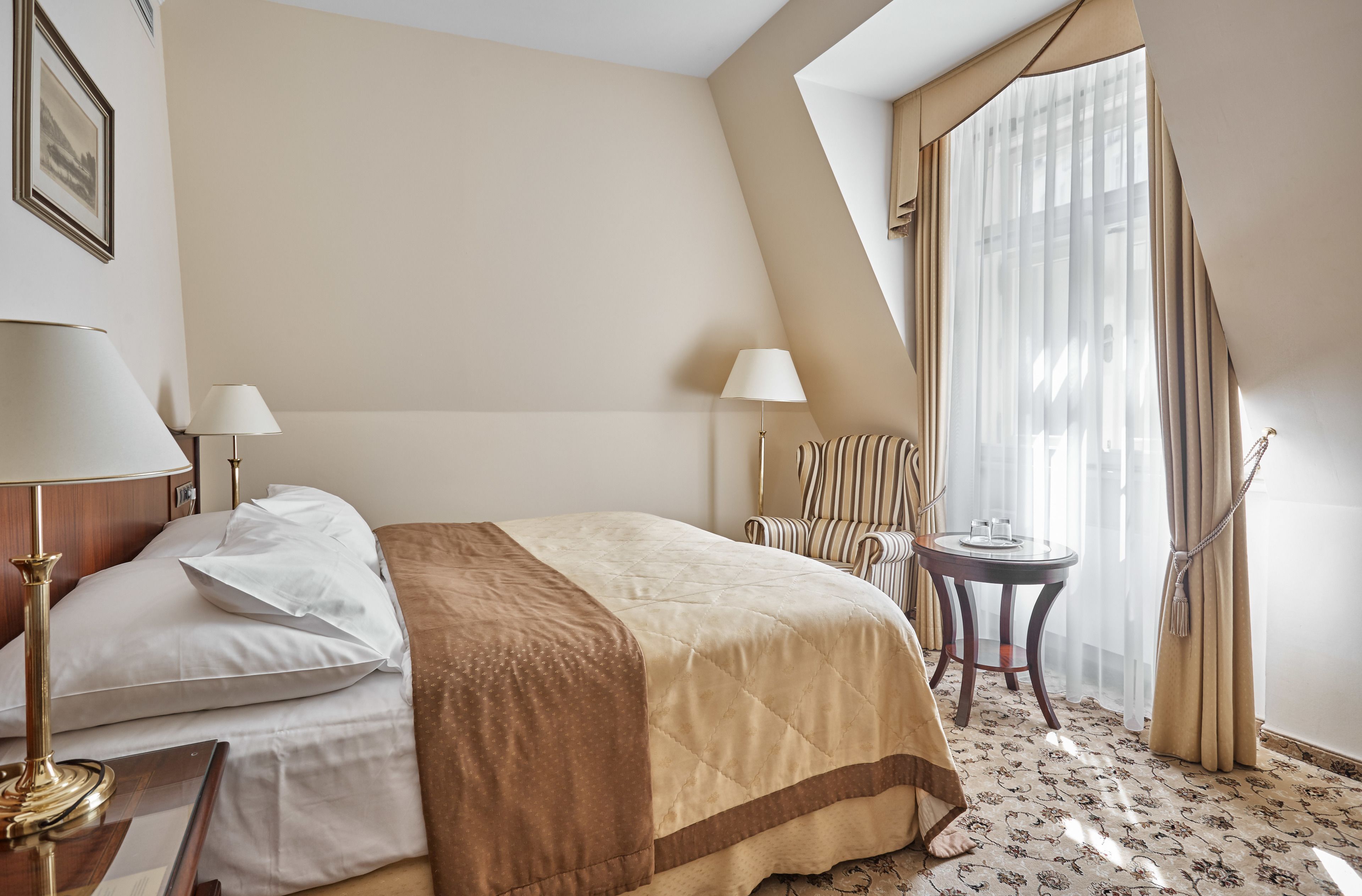 Habitación de hotel con cama, dos mesitas de noche, una silla, una mesita pequeña y una ventana con cortinas.