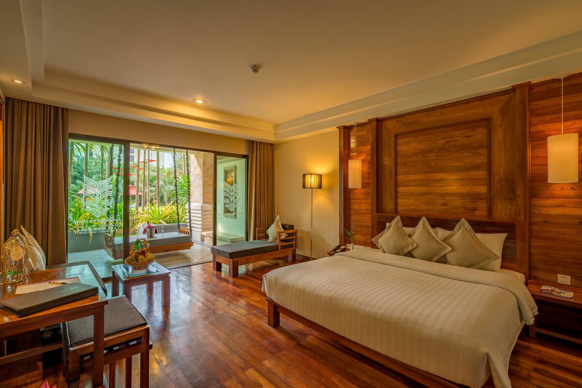 A spacious bedroom with a large bed, wooden headboard, and a view of lush greenery through the sliding glass door.