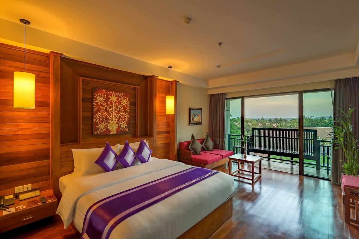 A hotel room with a large bed, wooden walls, a balcony with a view, and a potted plant.