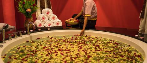 A large circular pool filled with water and flower petals, a towel rack with folded towels, and a vase with flowers.