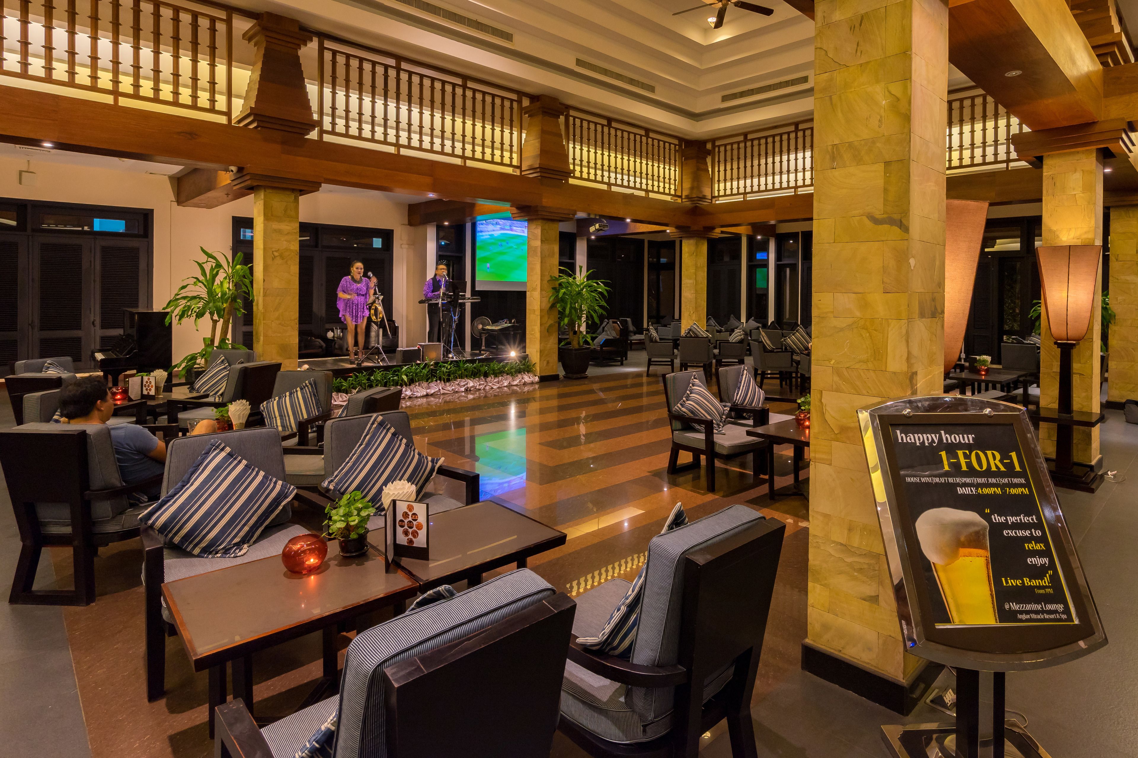 A spacious lobby with comfortable seating, a stage for live performances, and a sign promoting happy hour specials.