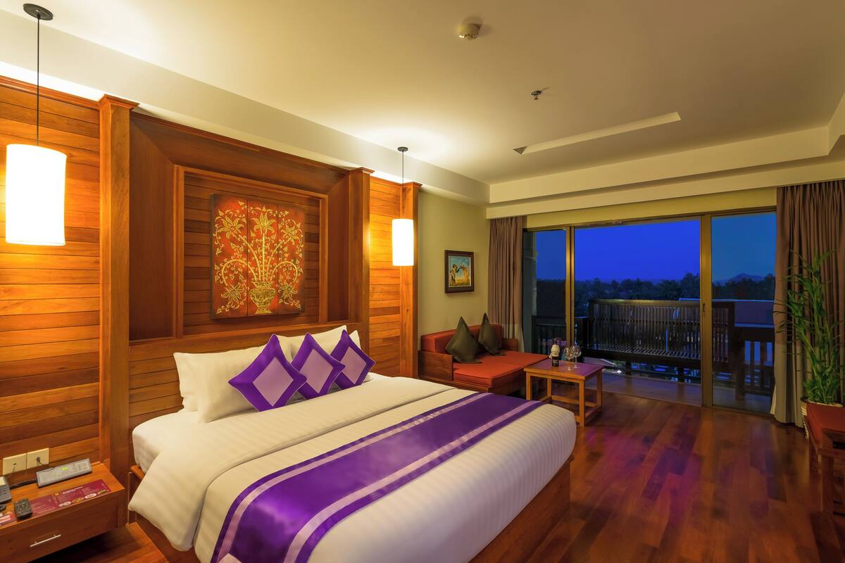 A hotel room with a large bed, a small table, a sofa, and a balcony with a view.