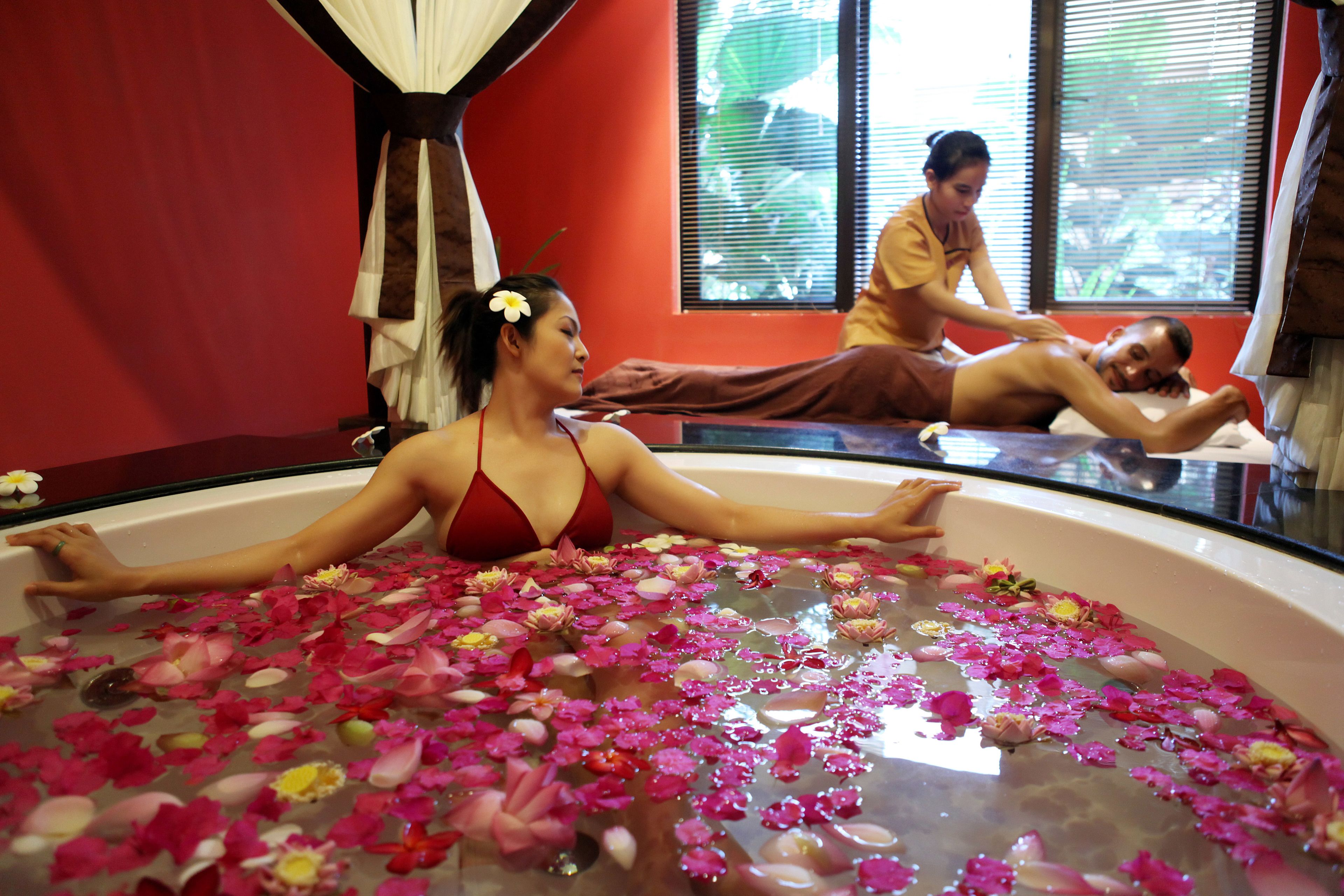A room with a bathtub filled with water and rose petals, a red wall, and a window with blinds.