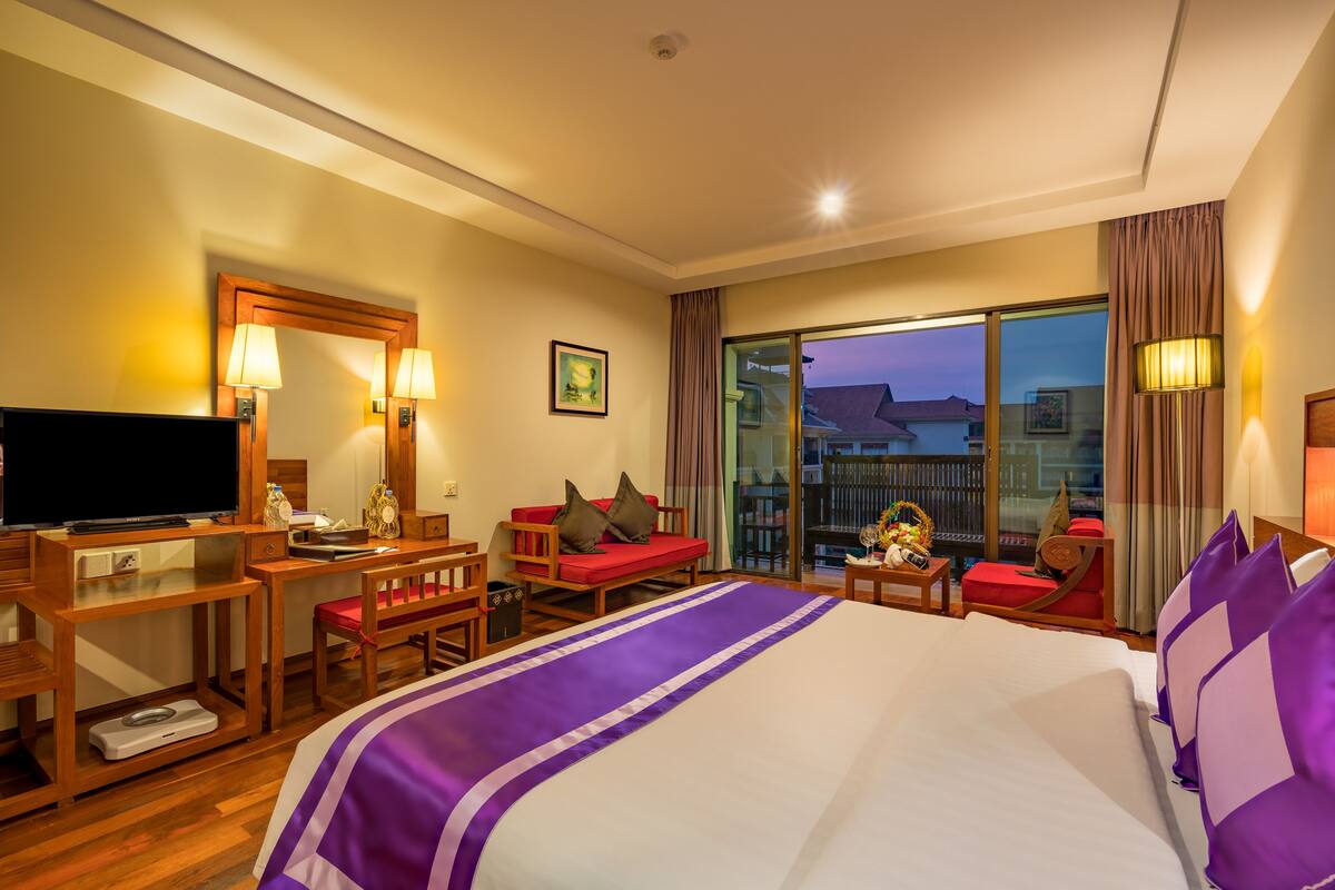 A hotel room with a large bed, a desk with a television, a seating area with a red sofa, and a balcony with a view of buildings.