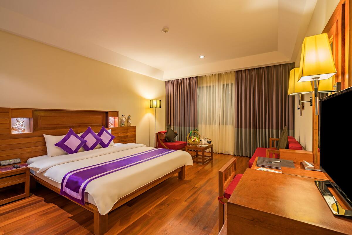 A hotel room with a large bed, wooden headboard, bedside tables, a desk with a chair, a television, and a window with curtains.