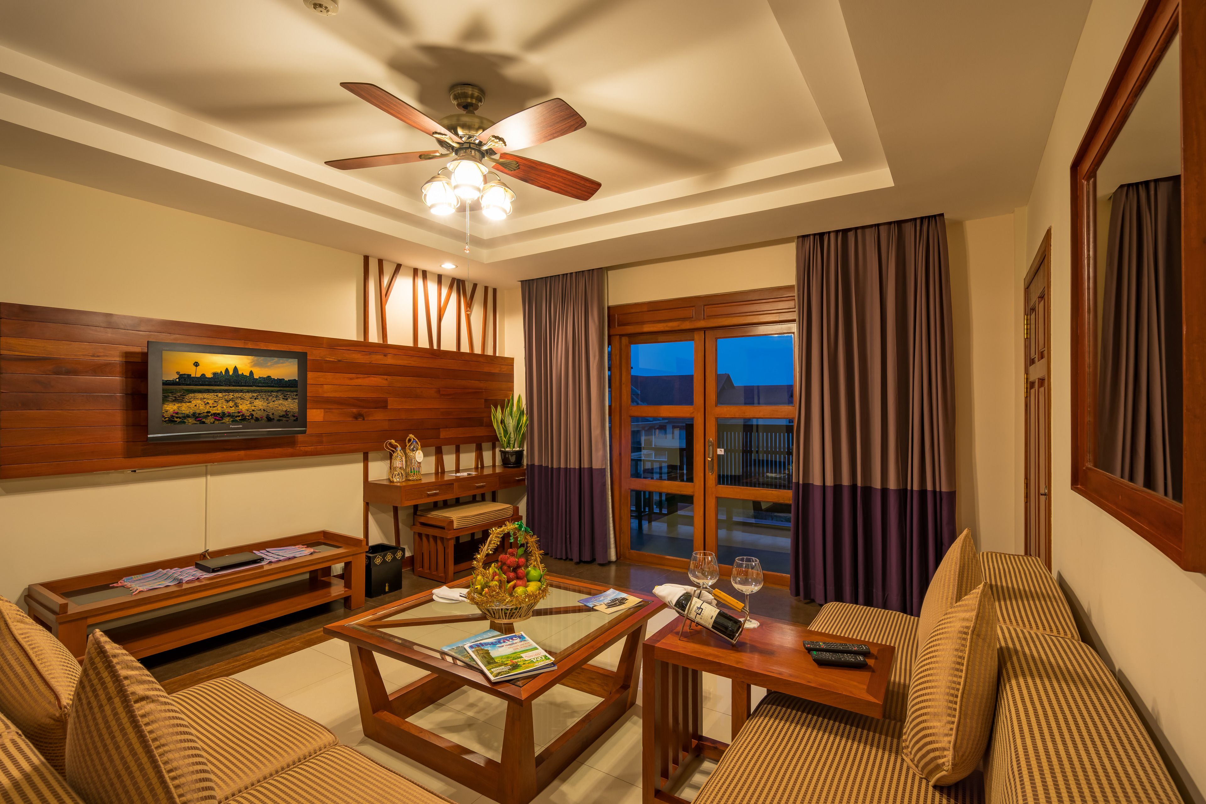 Un salon bien éclairé, doté d’un ventilateur de plafond, d’un canapé, d’une table basse avec des magazines, d’un téléviseur fixé au mur et d’un balcon avec des rideaux.