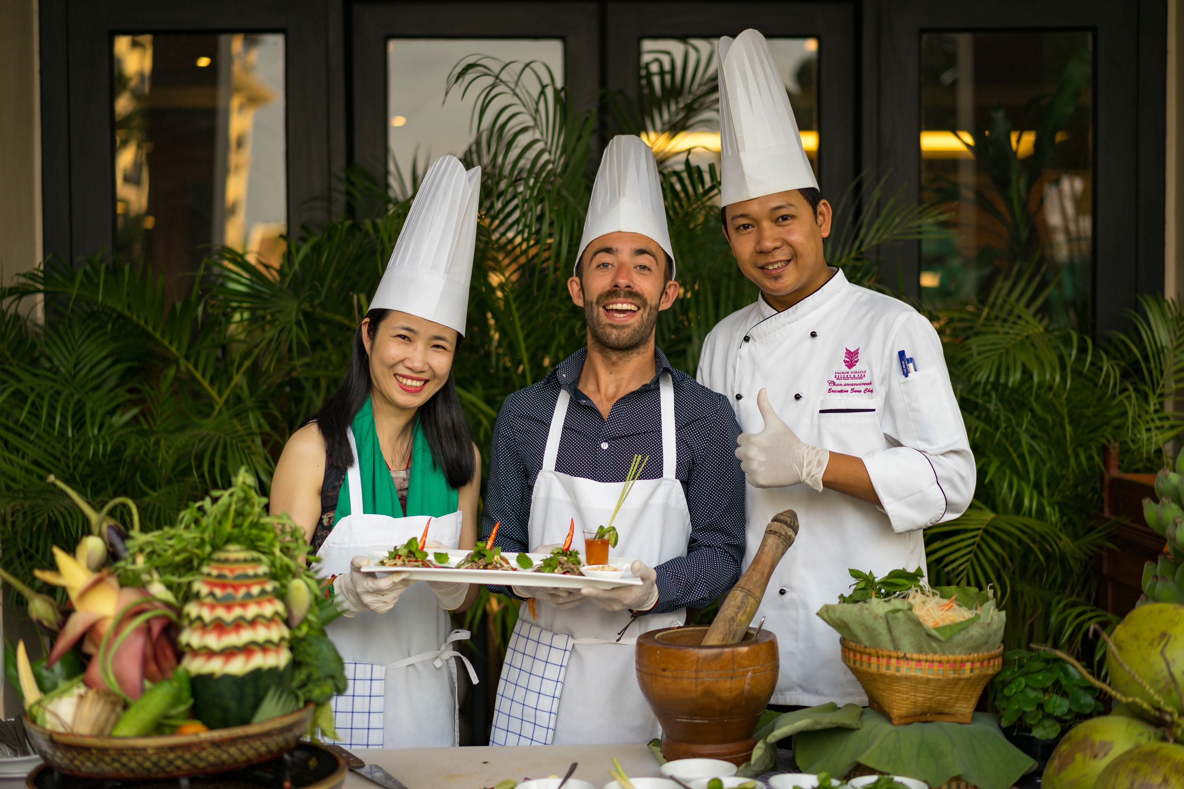 Three chefs in chef hats holding plates of food, with a variety of fresh vegetables and a mortar and pestle on a table.
