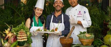 Three chefs in chef hats holding plates of food, with a variety of fresh vegetables and a mortar and pestle on a table.