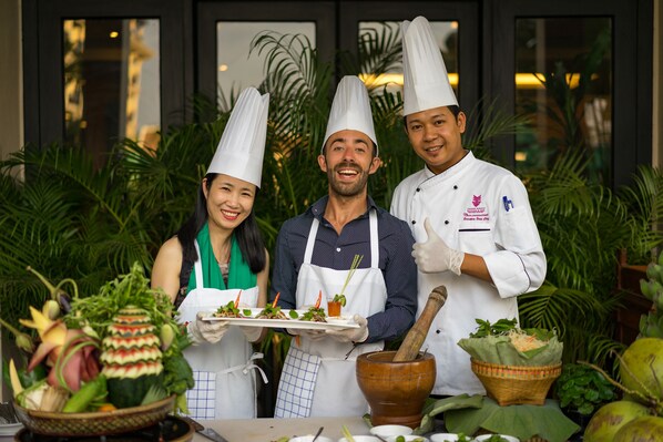 Tres cocineros con gorro de chef sostienen platos de comida, con una variedad de verduras frescas y un mortero y pilón sobre una mesa.