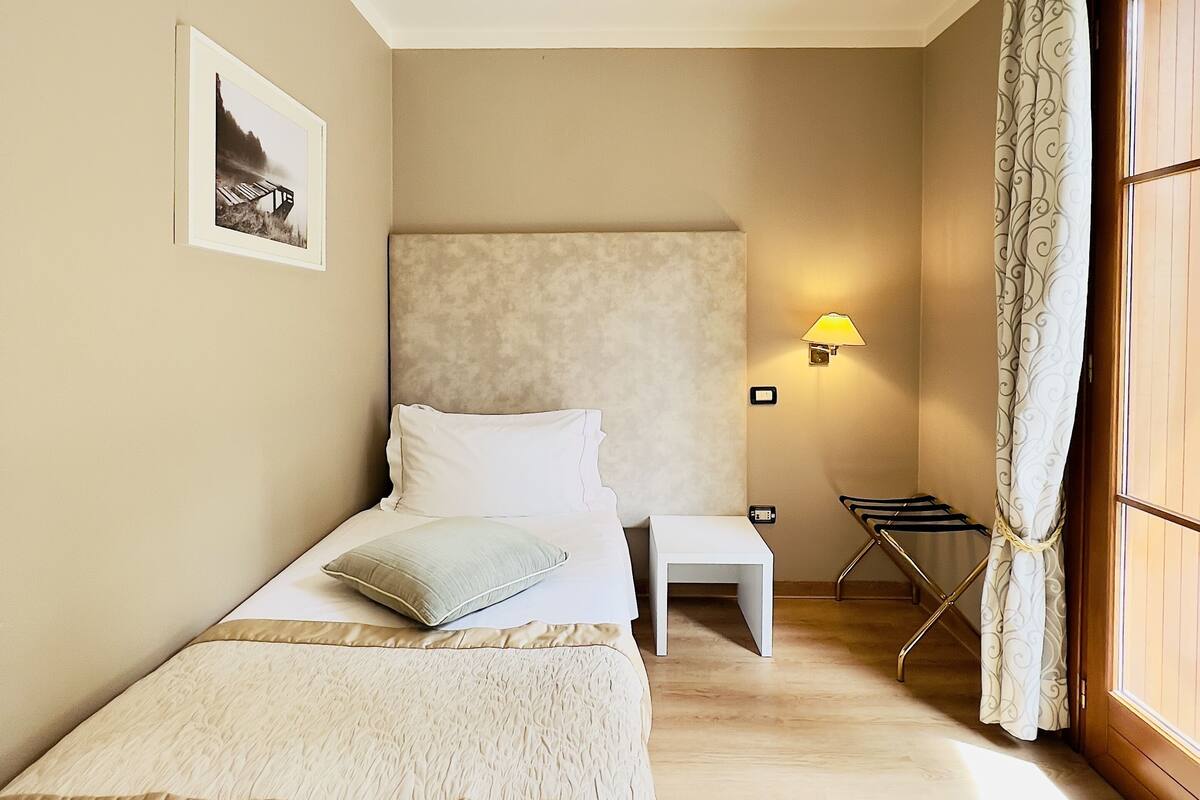 Une chambre avec deux lits, une tête de lit, une table de chevet, une lampe, un étendoir et un tableau au mur.