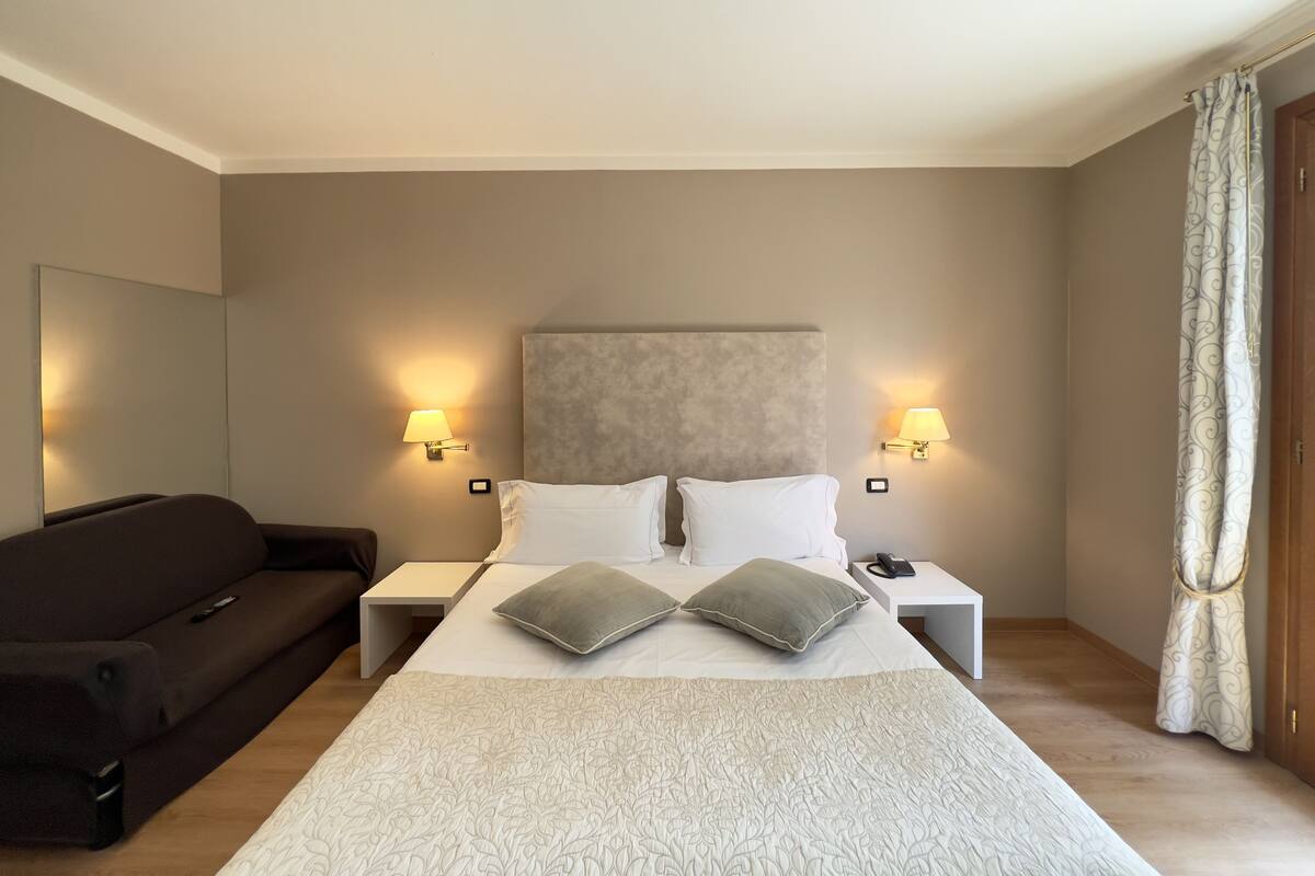 Une chambre d’hôtel avec un grand lit, deux tables de chevet, une petite table, un canapé et un rideau.