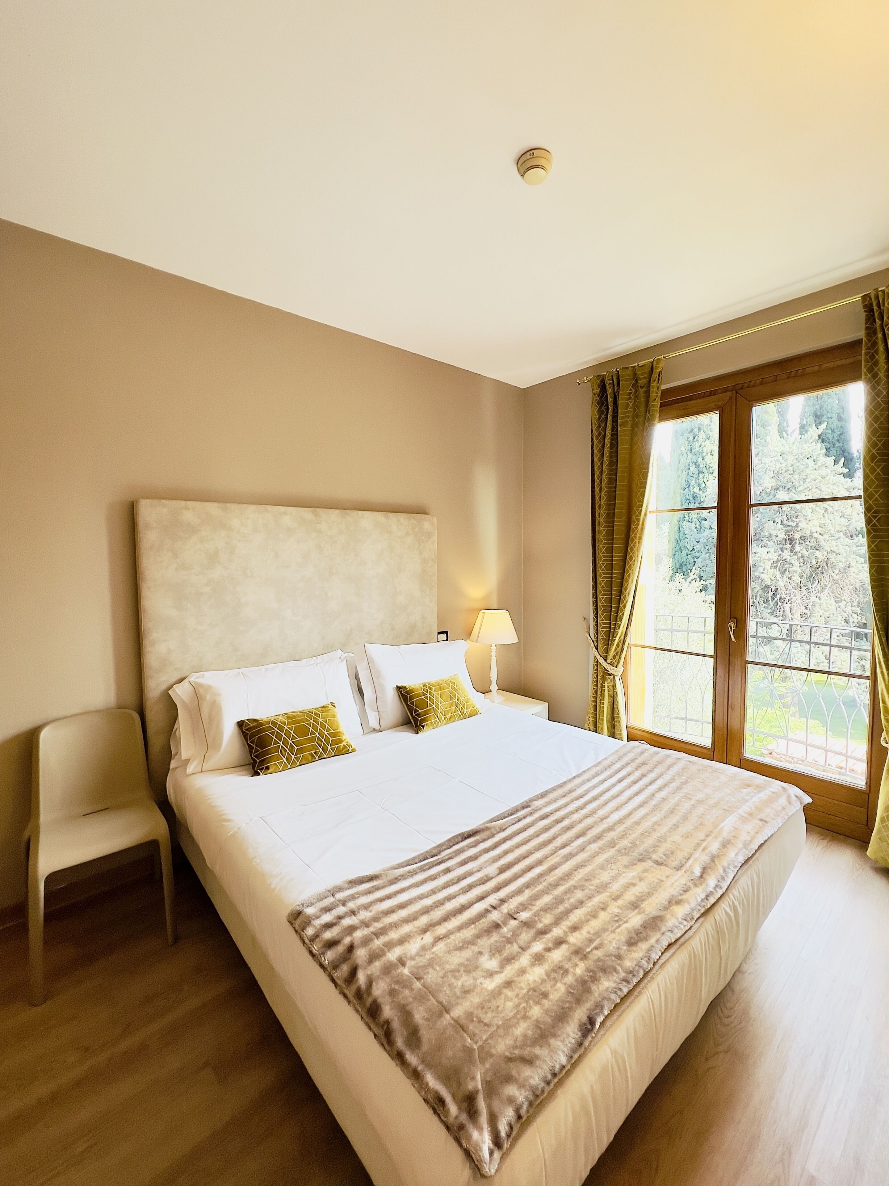 Habitación de hotel con una cama grande, una mesita de noche con lámpara, una silla y una ventana con cortinas.