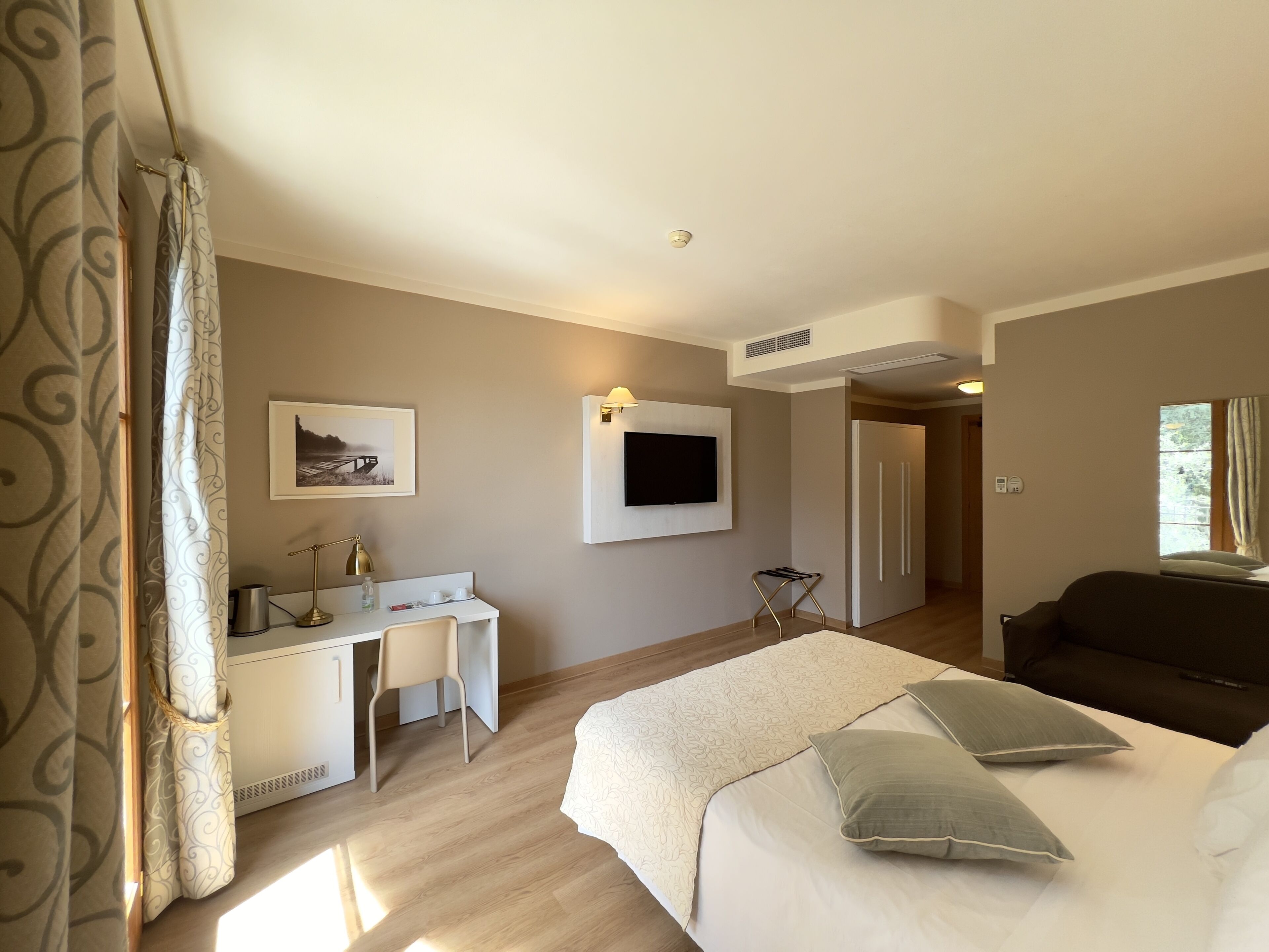 A hotel room with a bed, a desk with a chair, a television mounted on the wall, and a window.