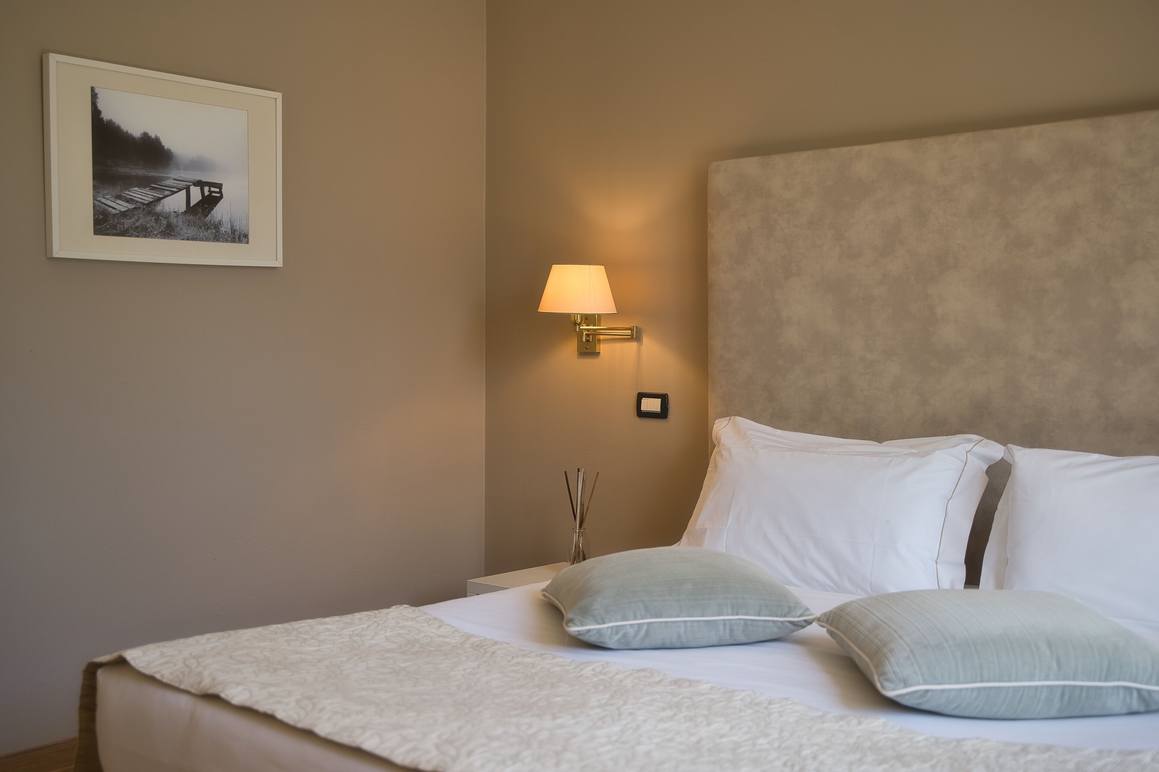 A neatly made bed with white linens and two pillows, a wall-mounted lamp, and a framed picture on the wall.