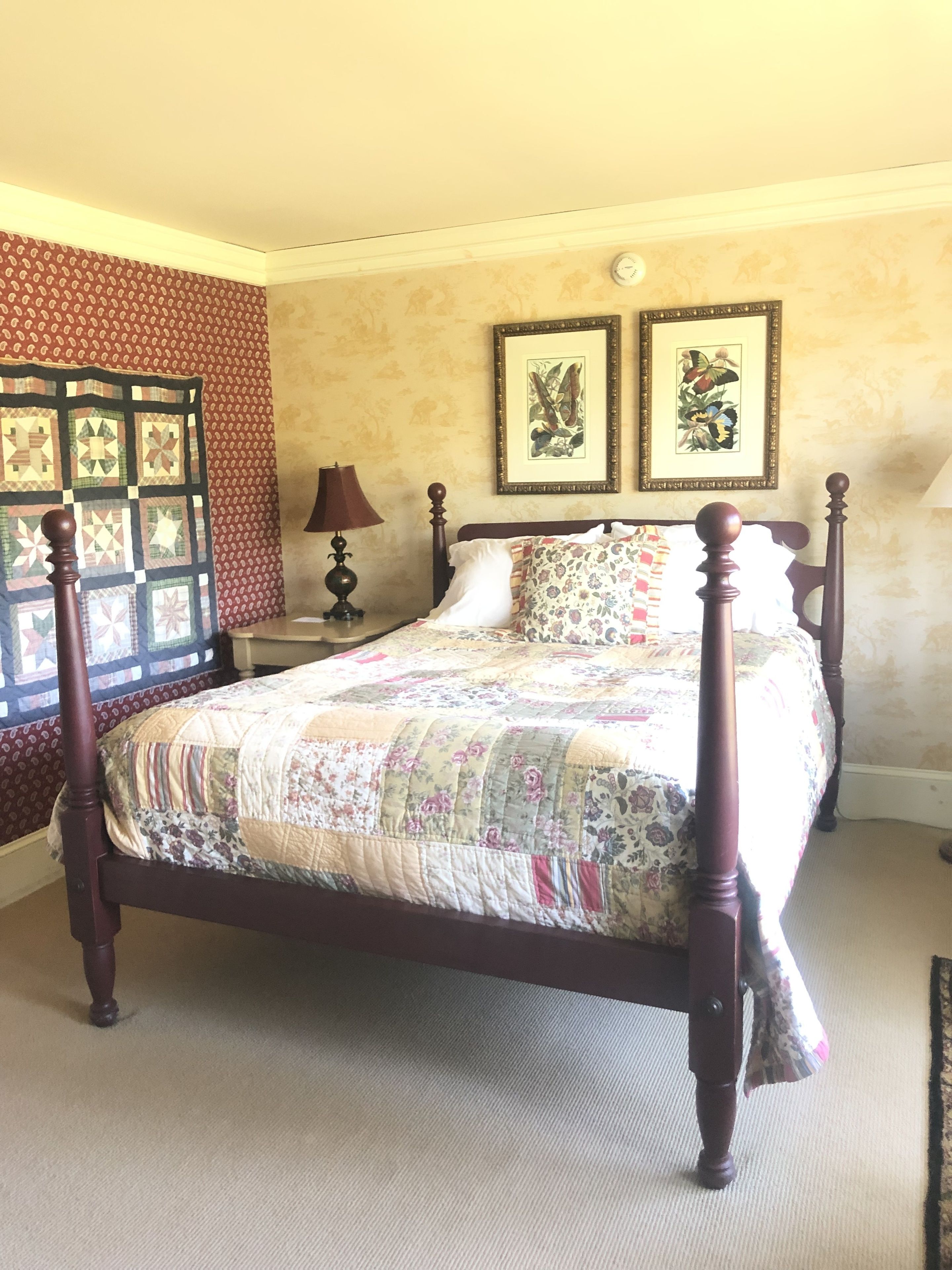 A four-poster bed with a floral quilt, a nightstand with a lamp, and two framed botanical prints on the wall.
