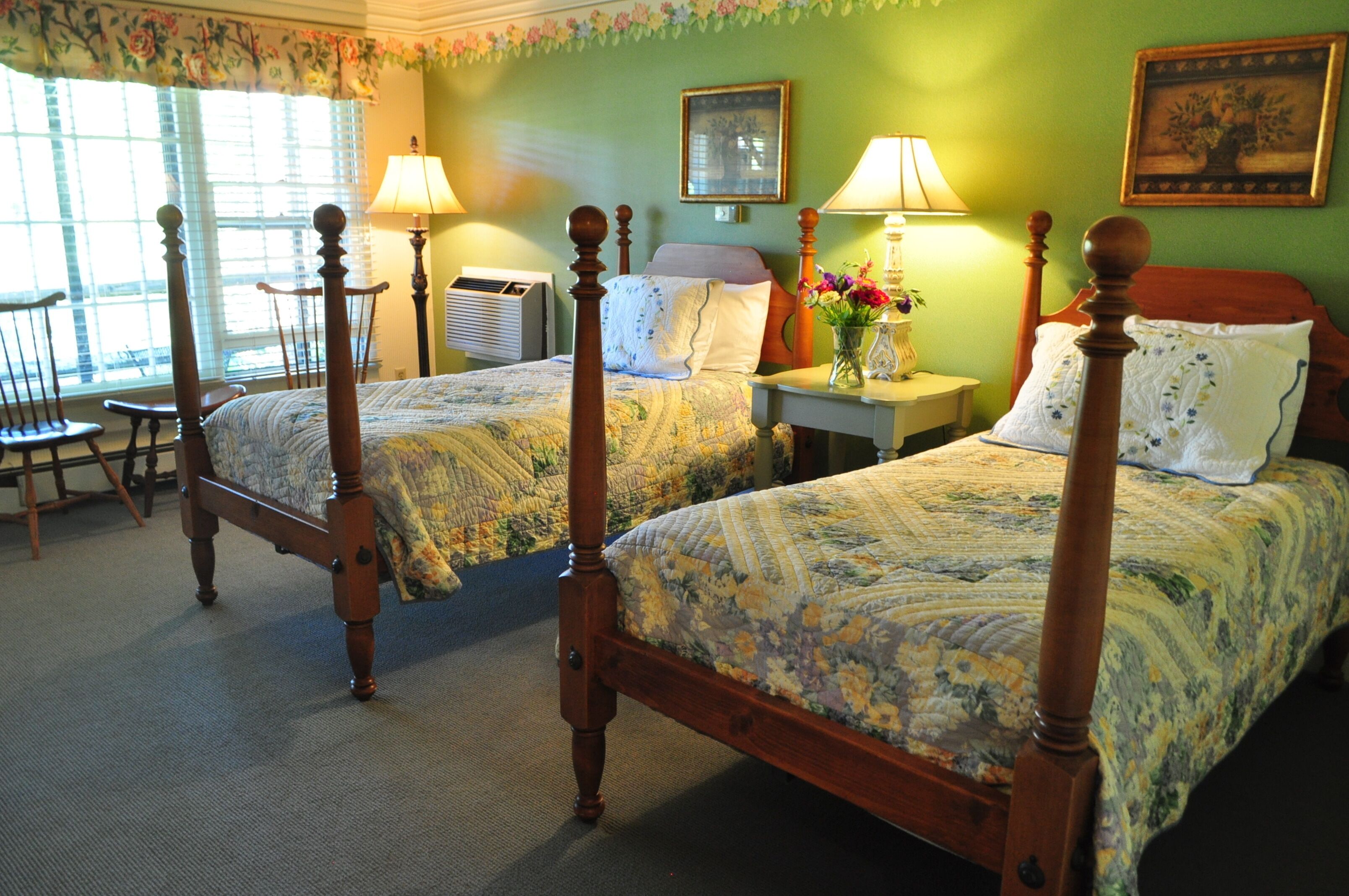 A room with two wooden four-poster beds, a nightstand with a lamp, a chair, and a window with blinds.