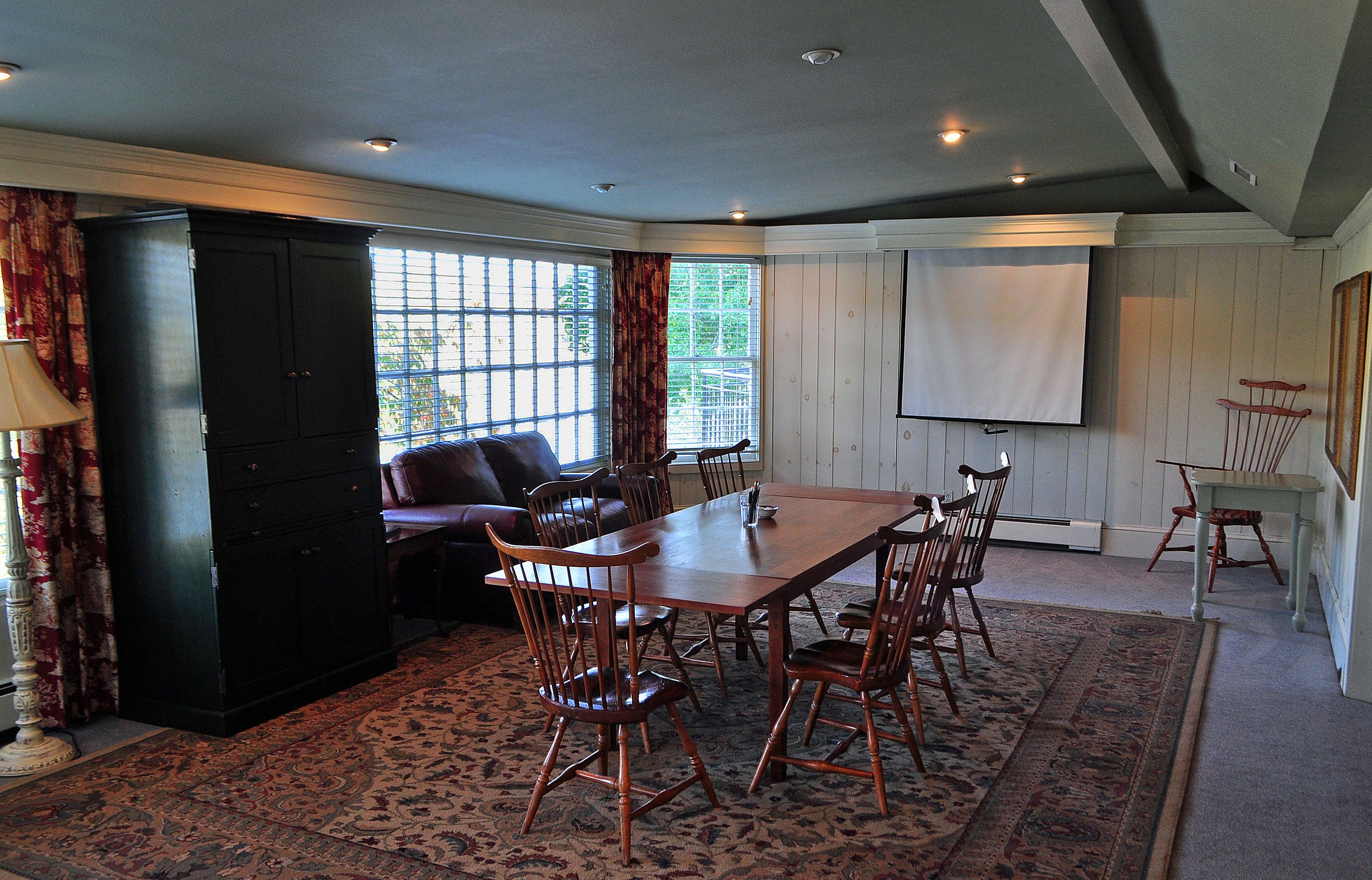 A dining area with a large wooden table, six chairs, a sofa, and a projector screen.