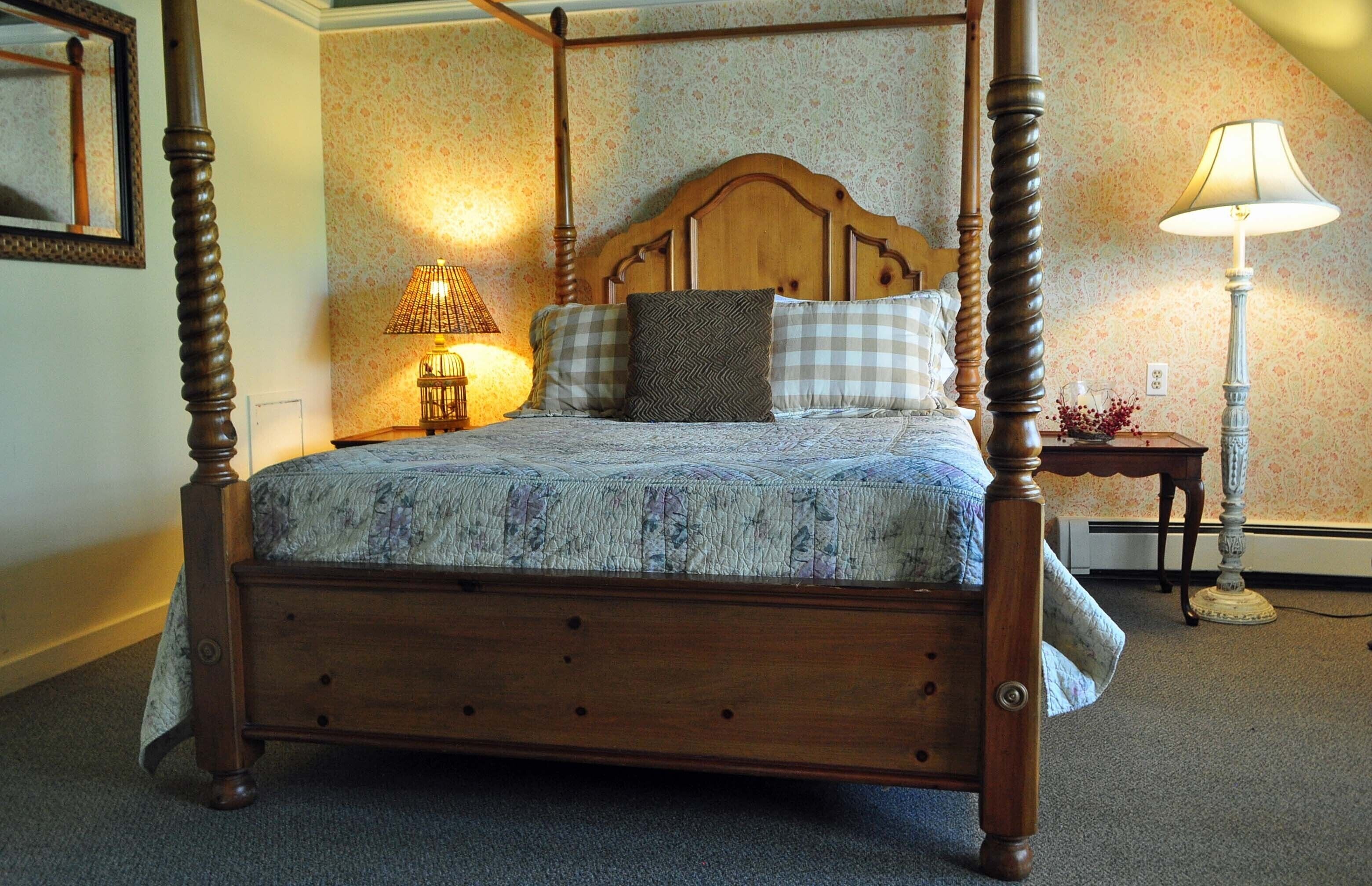 A four-poster bed with a floral-patterned bedspread, a nightstand with a lamp, and a mirror on the wall.