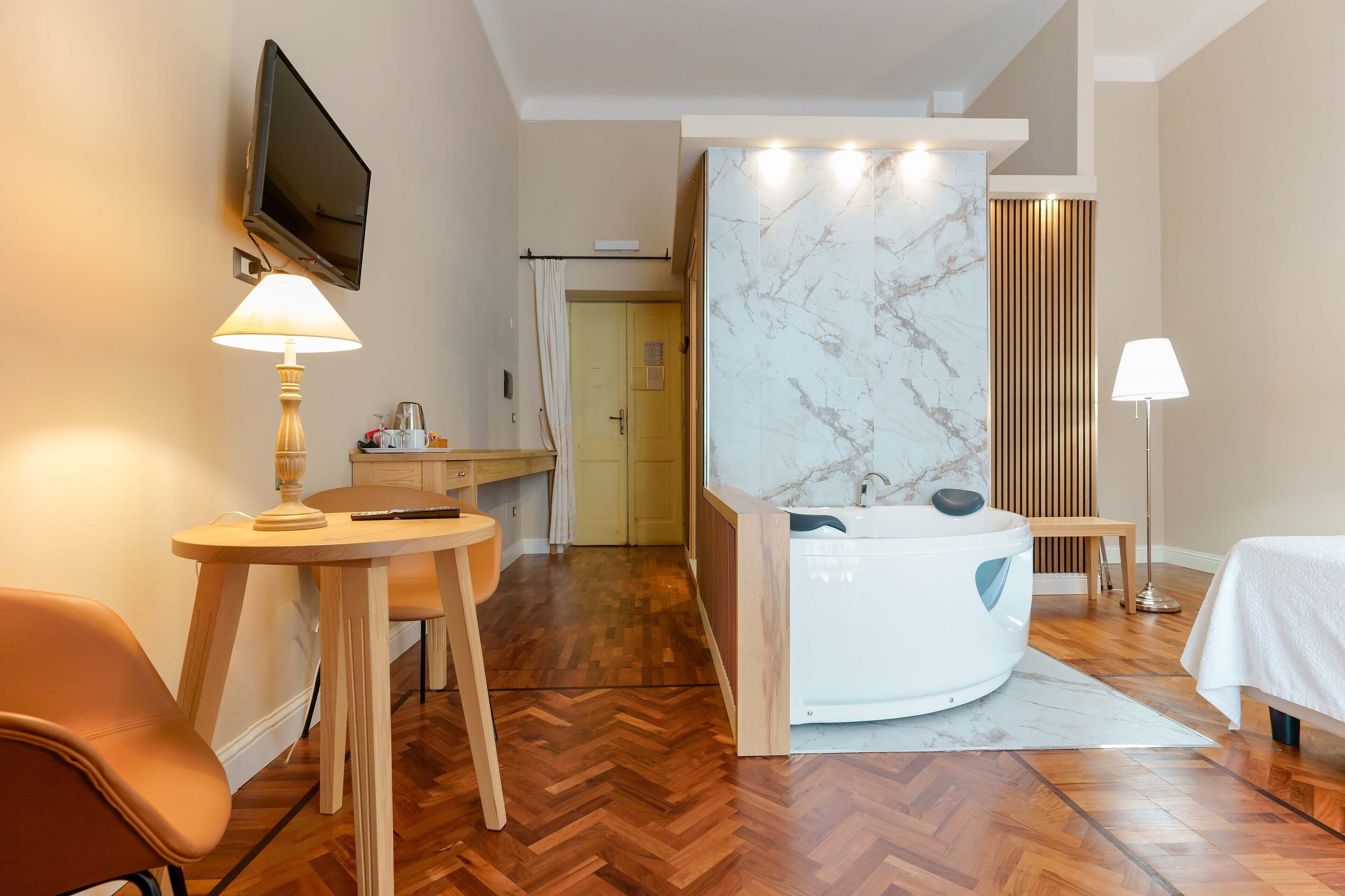 Une chambre d’hôtel avec un plancher en bois, un téléviseur fixé au mur, un bureau avec une lampe, un lit et une salle de bain avec un mur en marbre et une baignoire.