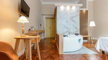 Une chambre d’hôtel avec un plancher en bois, un téléviseur fixé au mur, un bureau avec une lampe, un lit et une salle de bain avec un mur en marbre et une baignoire.