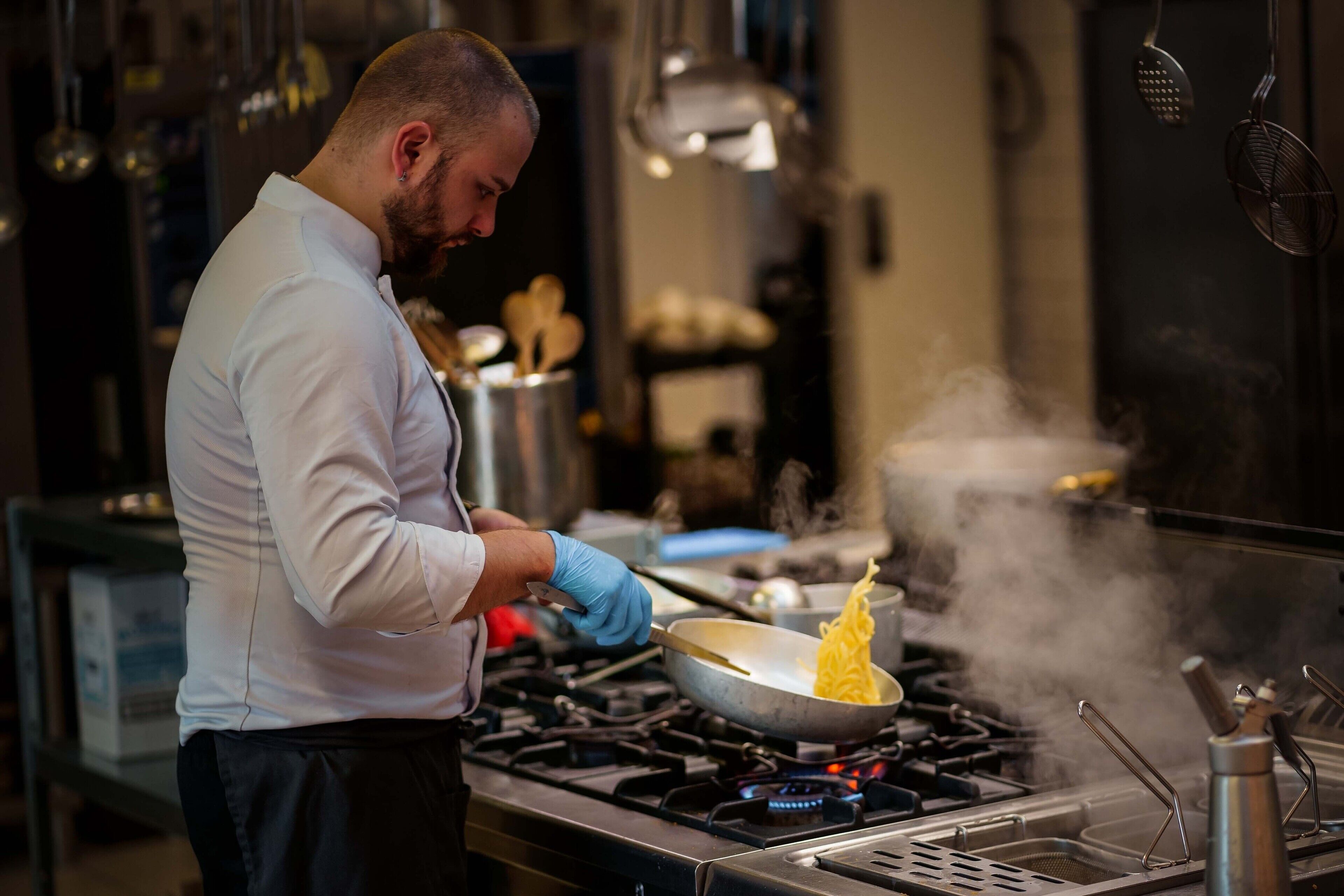 A chef cooking with a pan on a stove, steam rising, and kitchen utensils in the background.