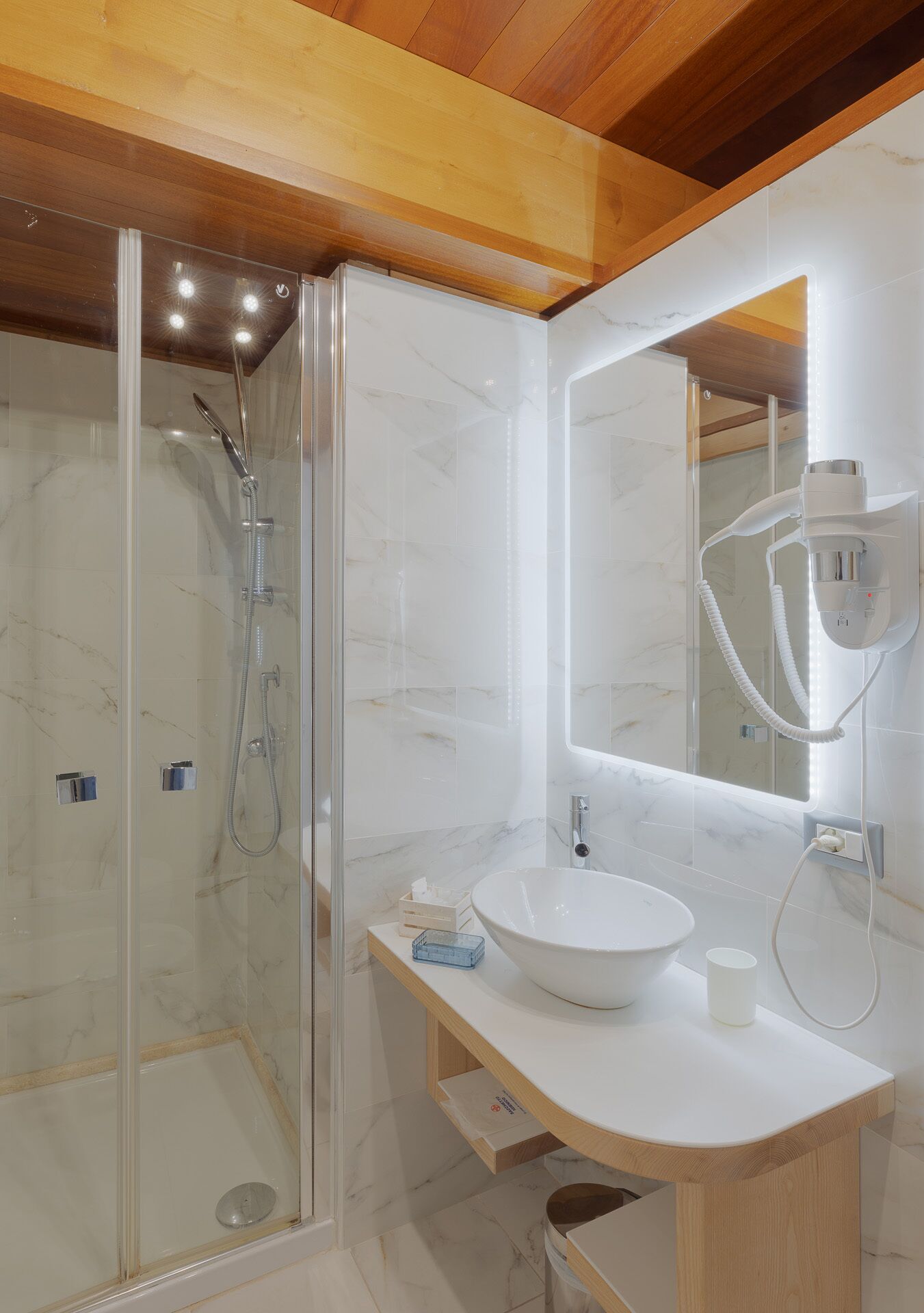 Un baño moderno con ducha de vidrio, lavamanos blanco e iluminación integrada en el espejo.