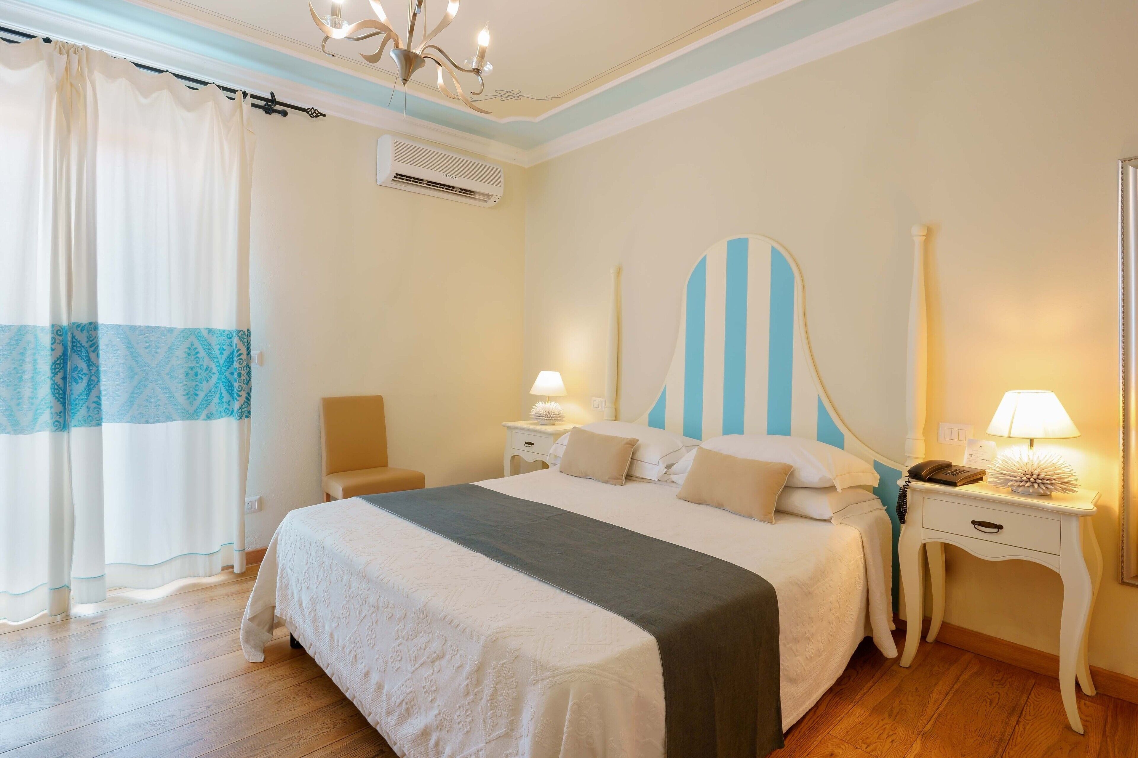 Une chambre à coucher avec un grand lit, deux tables de chevet, une commode, un lustre et une fenêtre avec des rideaux.