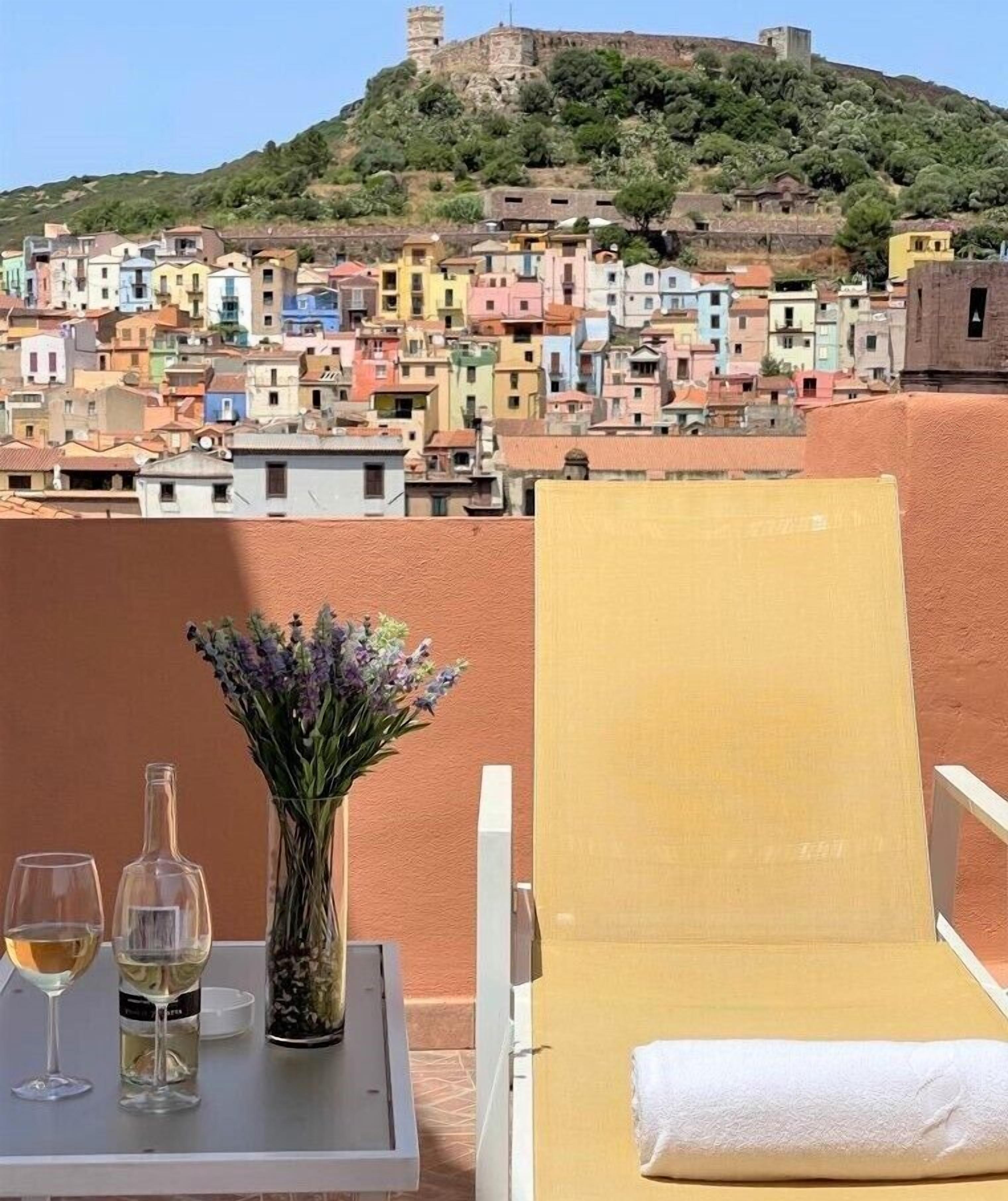 Ein Balkon mit Tisch, zwei Weingläsern und einer Blumenvase. Außerdem ist eine Sonnenliege zu sehen.