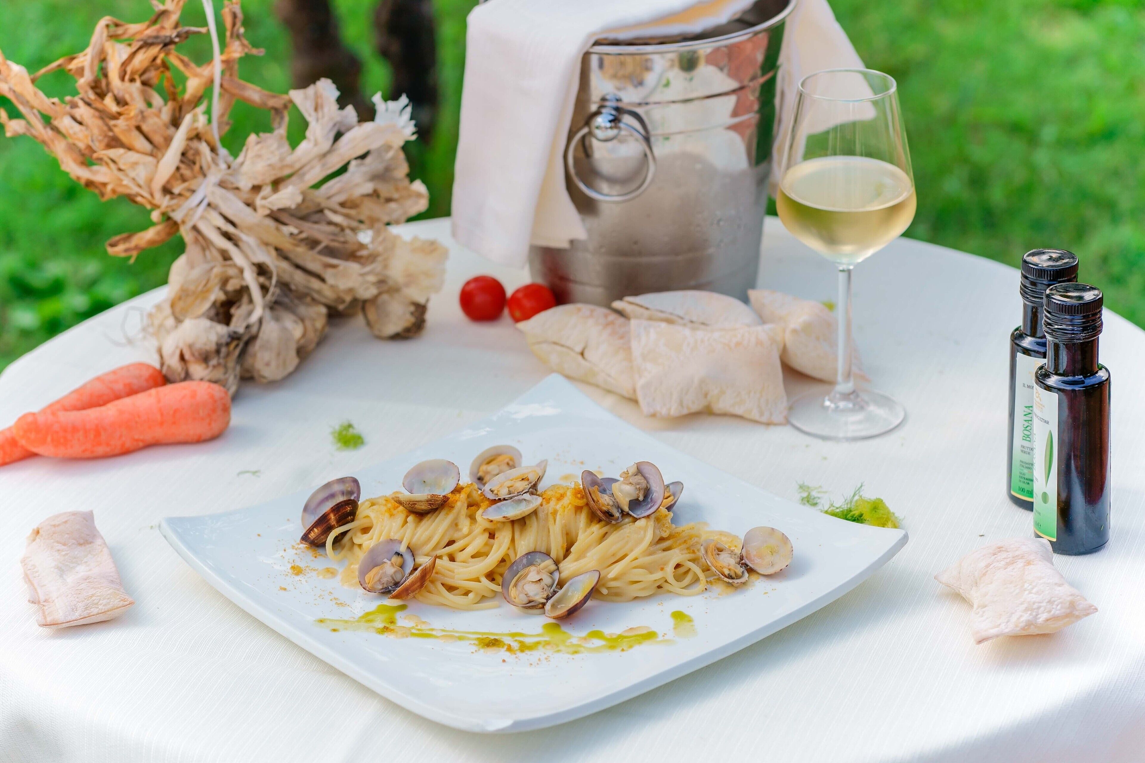 A dining table with a plate of pasta, clams, and vegetables, a wine glass, and a bottle of olive oil.