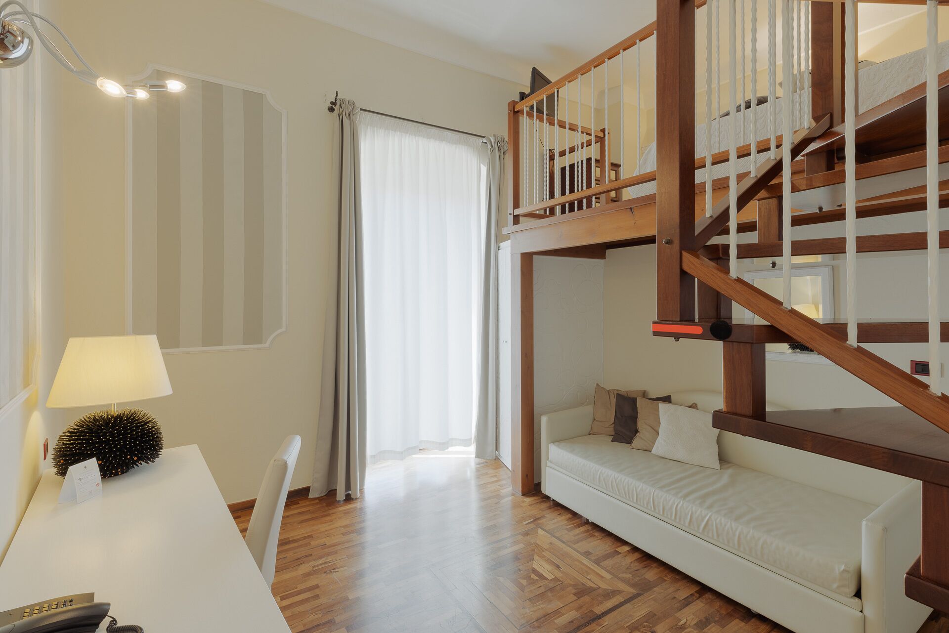 Una habitación con una escalera de madera, un sofá blanco, un escritorio con lámpara y un espejo.