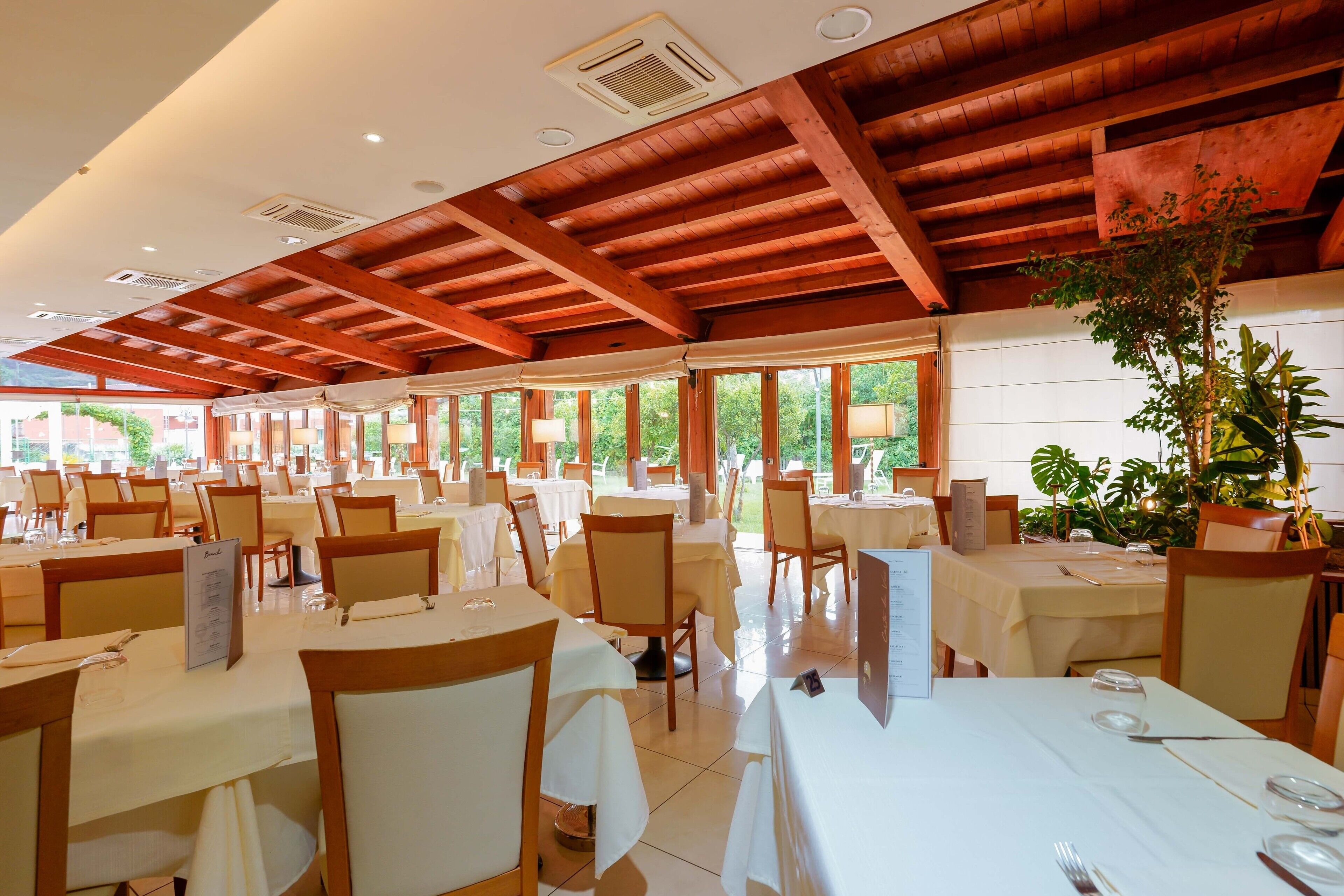 A spacious dining area with multiple tables and chairs, wooden ceiling beams, and large windows allowing natural light.