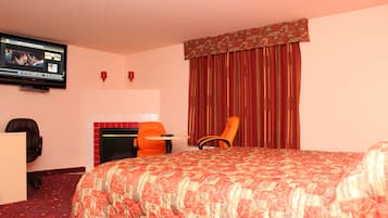 A hotel room with a large bed, a television mounted on the wall, a fireplace, and two chairs.
