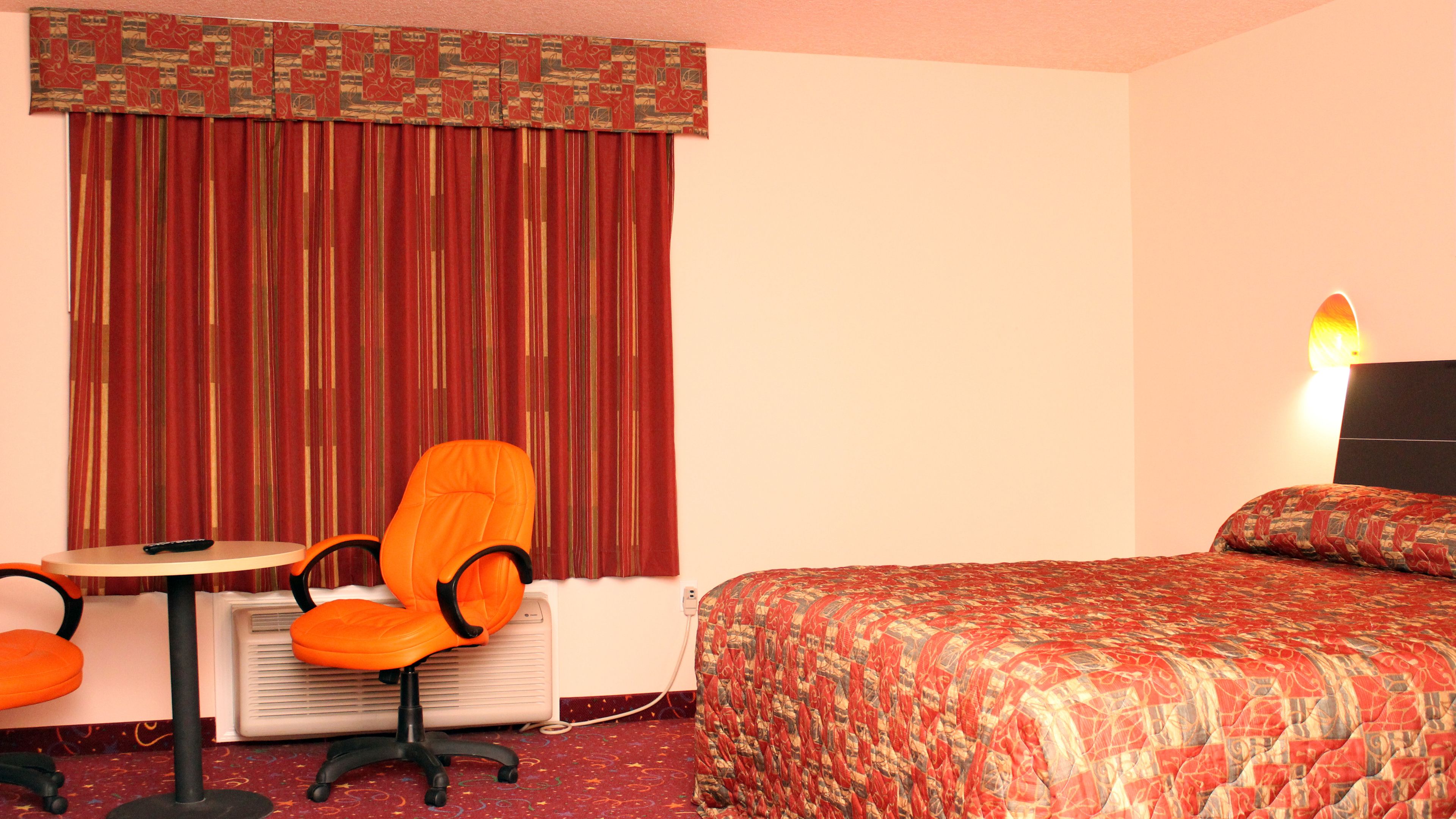A hotel room with a bed, an orange chair, a small table with a remote control, and a wall-mounted lamp.