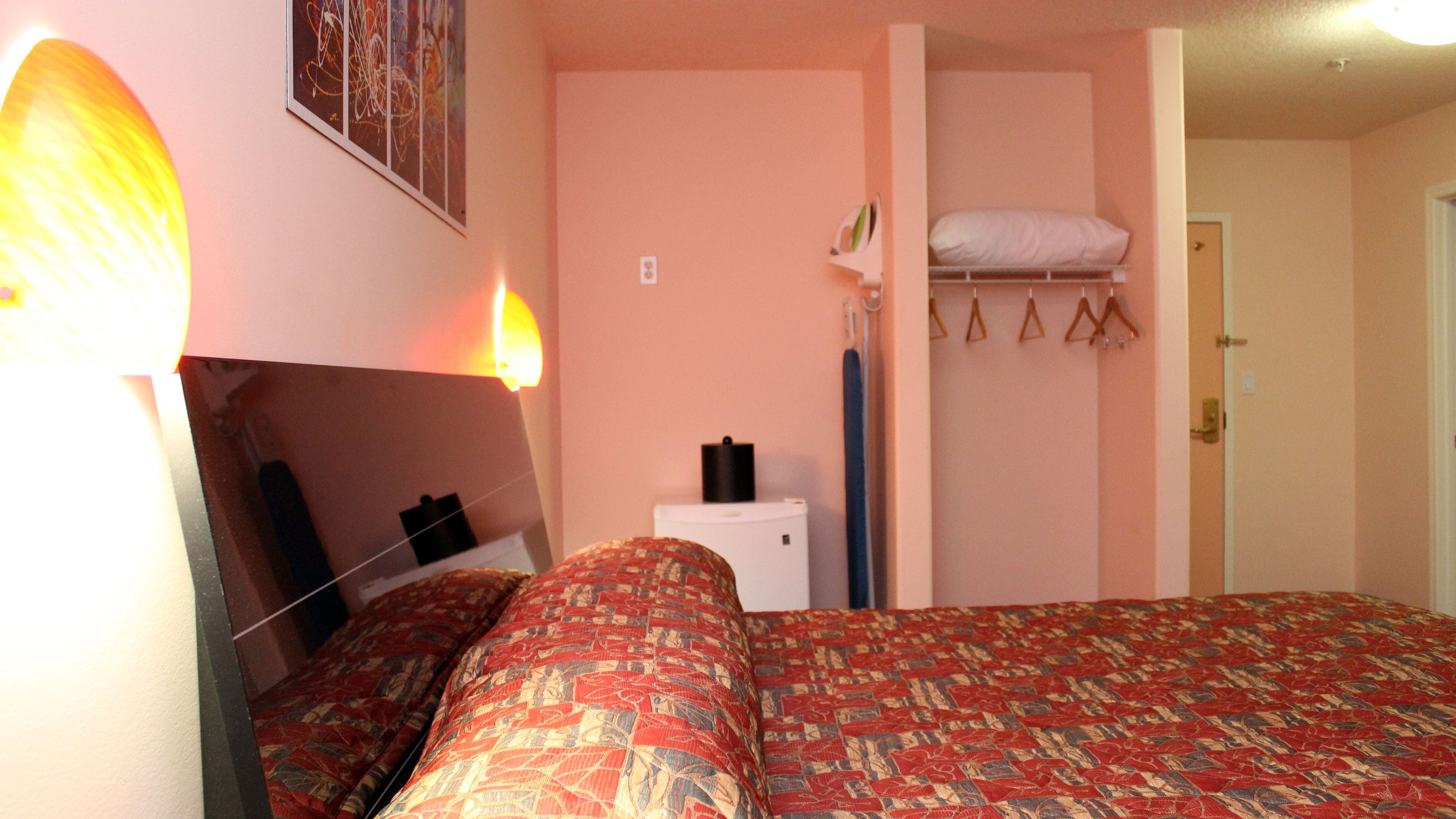 A bedroom with a bed, a bedside table, a hanging light, a closet with a hanging shelf, and a door.