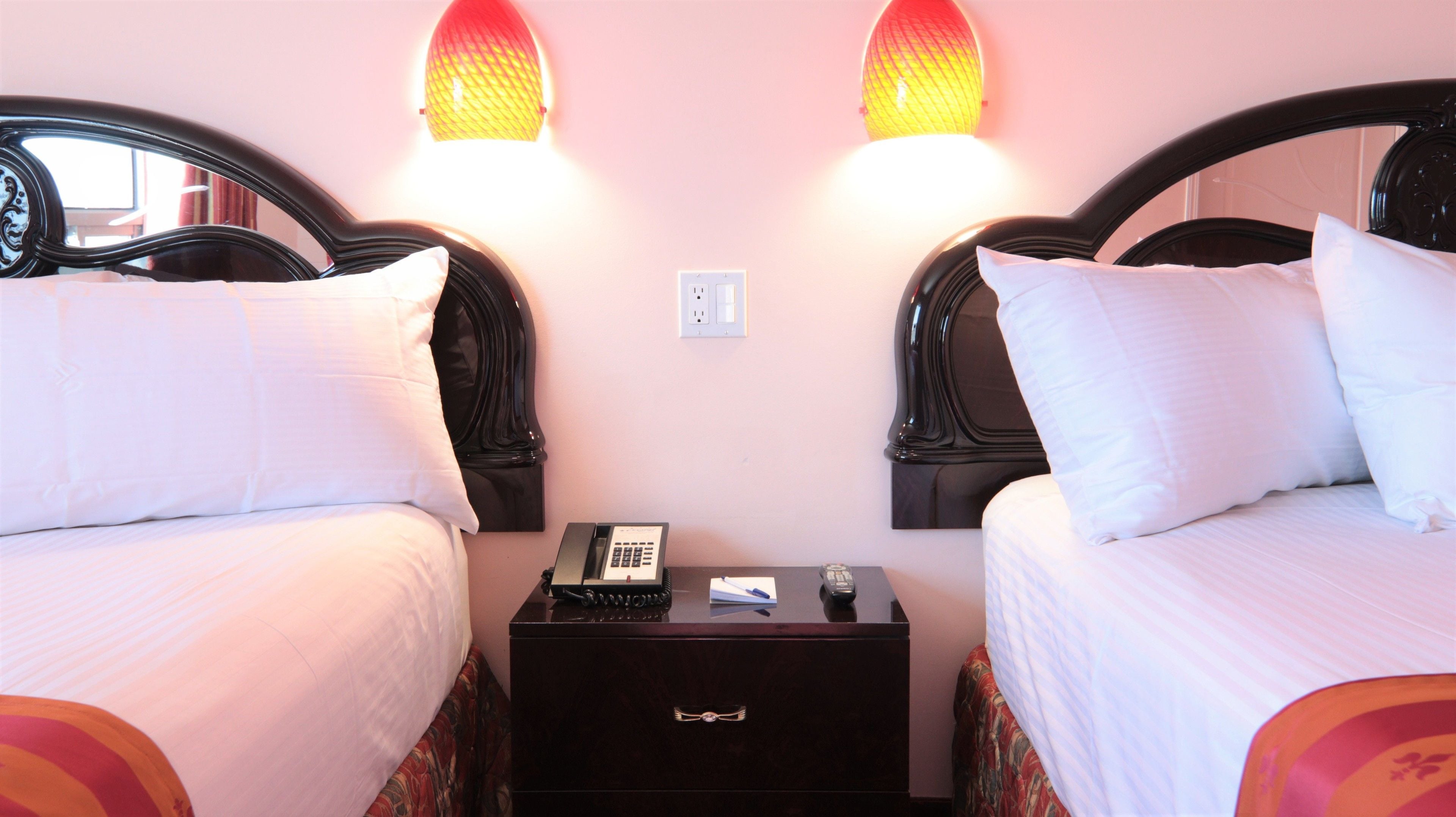 Une chambre d’hôtel avec deux lits, chacun doté de draps blancs et d’une tête de lit noire. Une table de chevet se trouve entre les deux lits, sur laquelle reposent un téléphone, un bloc-notes et un stylo. La chambre a un mur rose et une fenêtre avec des rideaux.