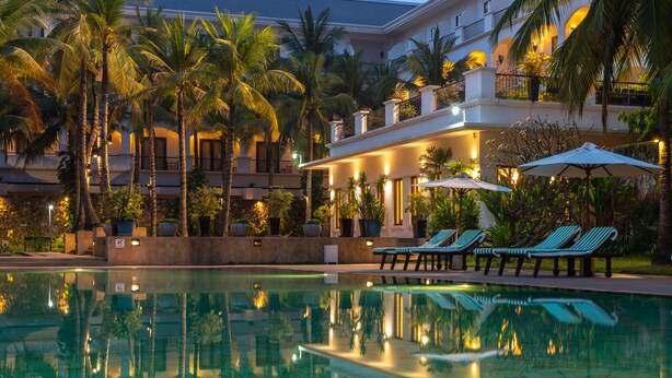 A swimming pool with lounge chairs and umbrellas, surrounded by palm trees and a building with balconies. - Lotus Blanc Resort (Siem Reap)
