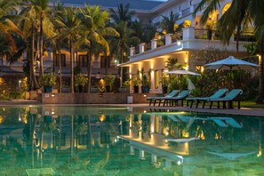 A swimming pool with lounge chairs and umbrellas, surrounded by palm trees and a building with balconies. - Lotus Blanc Resort (Siem Reap)