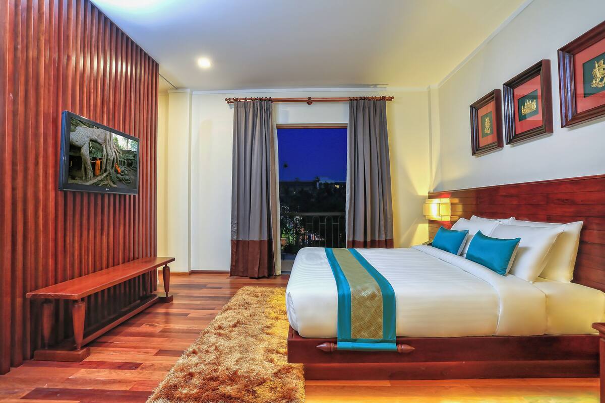 A hotel room with a large bed, wooden paneling, a bench, a view of the outside, and framed pictures on the wall.