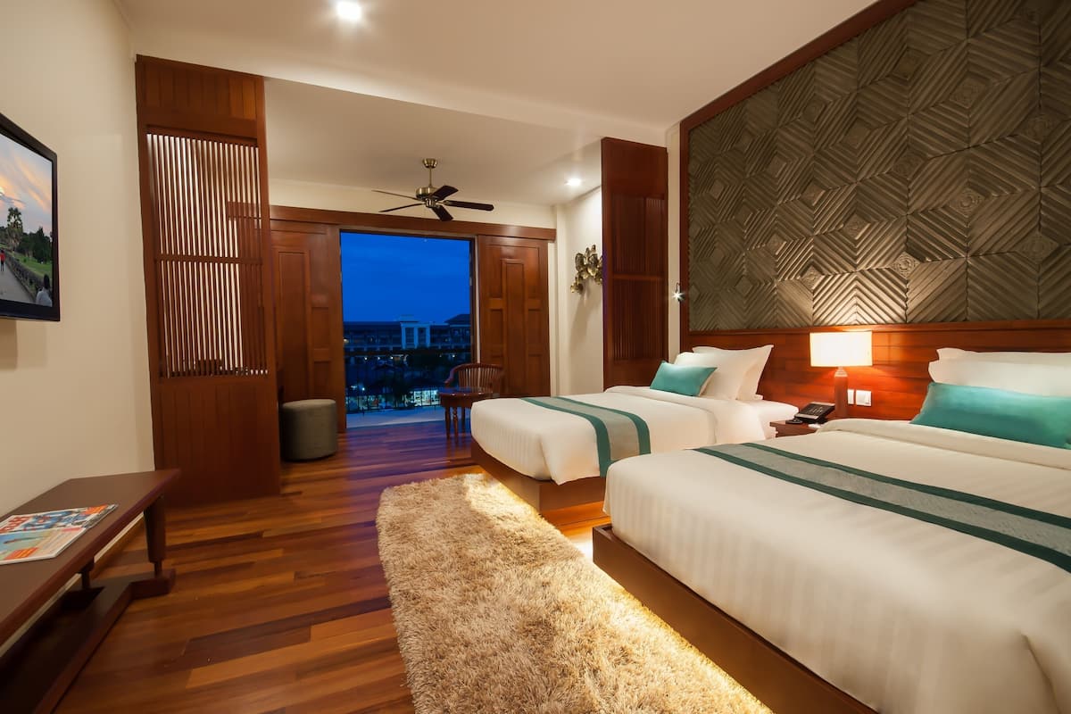 A hotel room with two beds, a wooden floor, a view of the city at dusk, a ceiling fan, and a nightstand with a lamp.