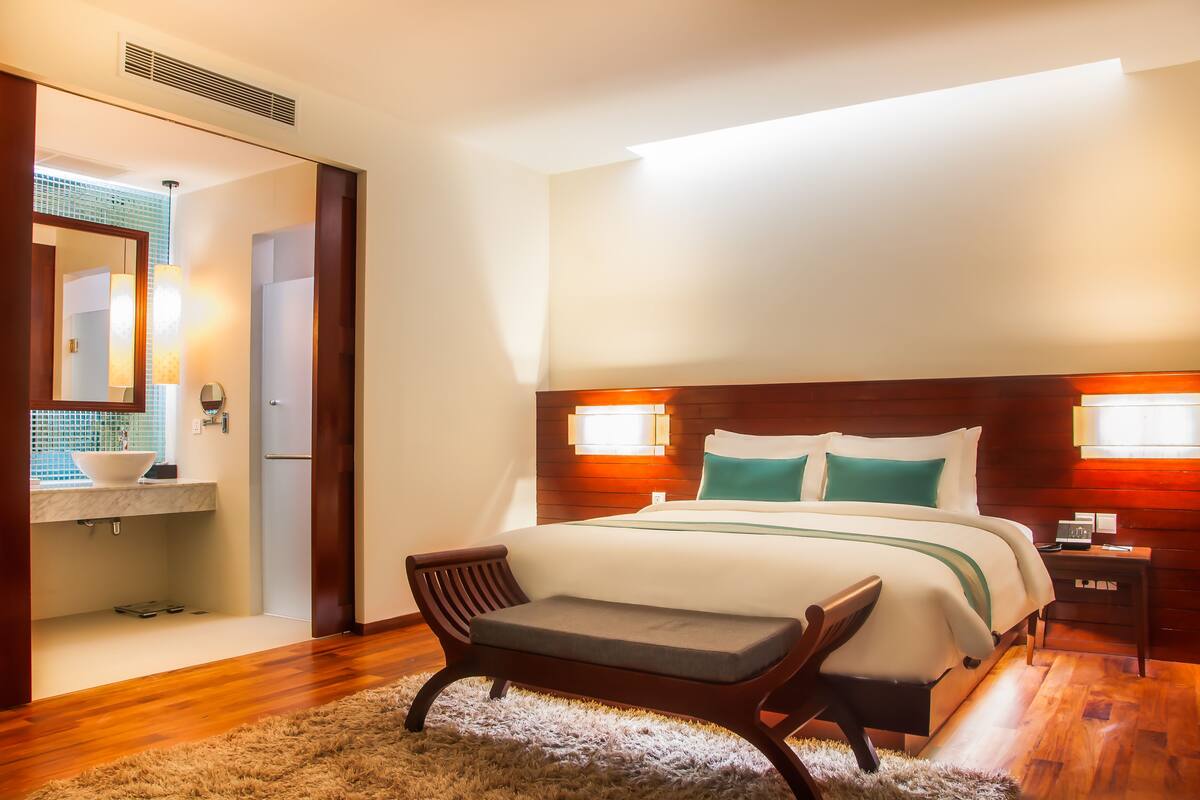 A hotel room with a large bed, bedside tables, a bench, a mirror, and a bathroom with a sink and mirror.
