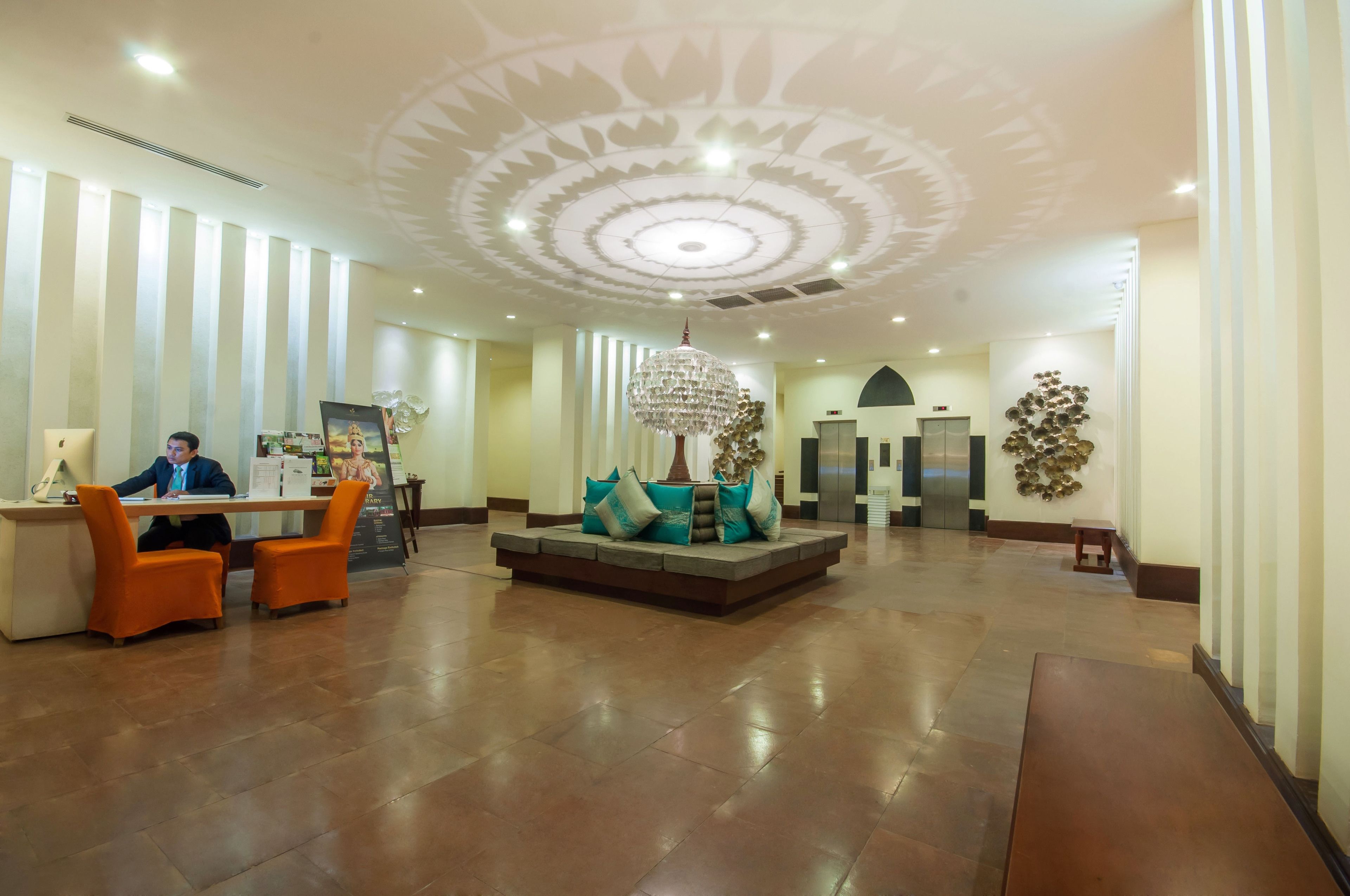 A spacious lobby with a reception desk, orange chairs, a central seating area with cushions, a chandelier, and a patterned ceiling.