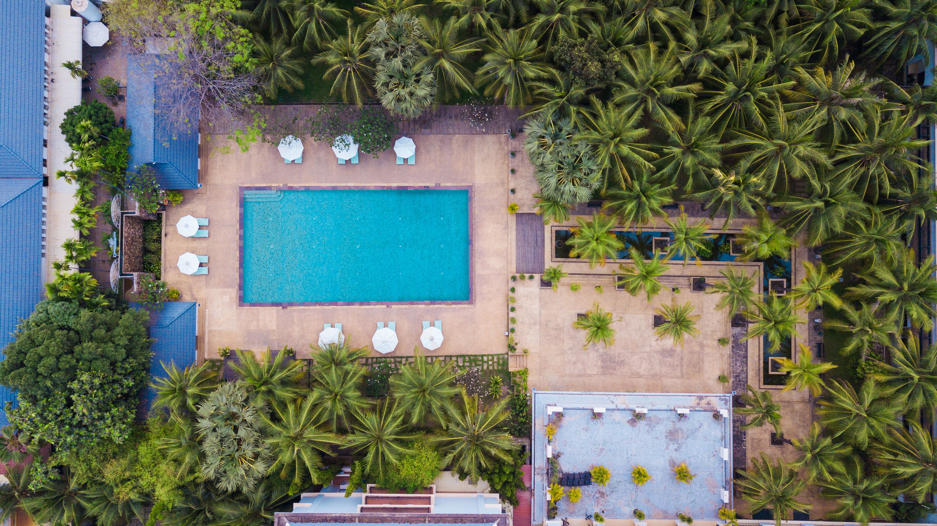 Vue aérienne d’un complexe hôtelier doté d’une piscine centrale, entourée de palmiers et d’espaces de détente.