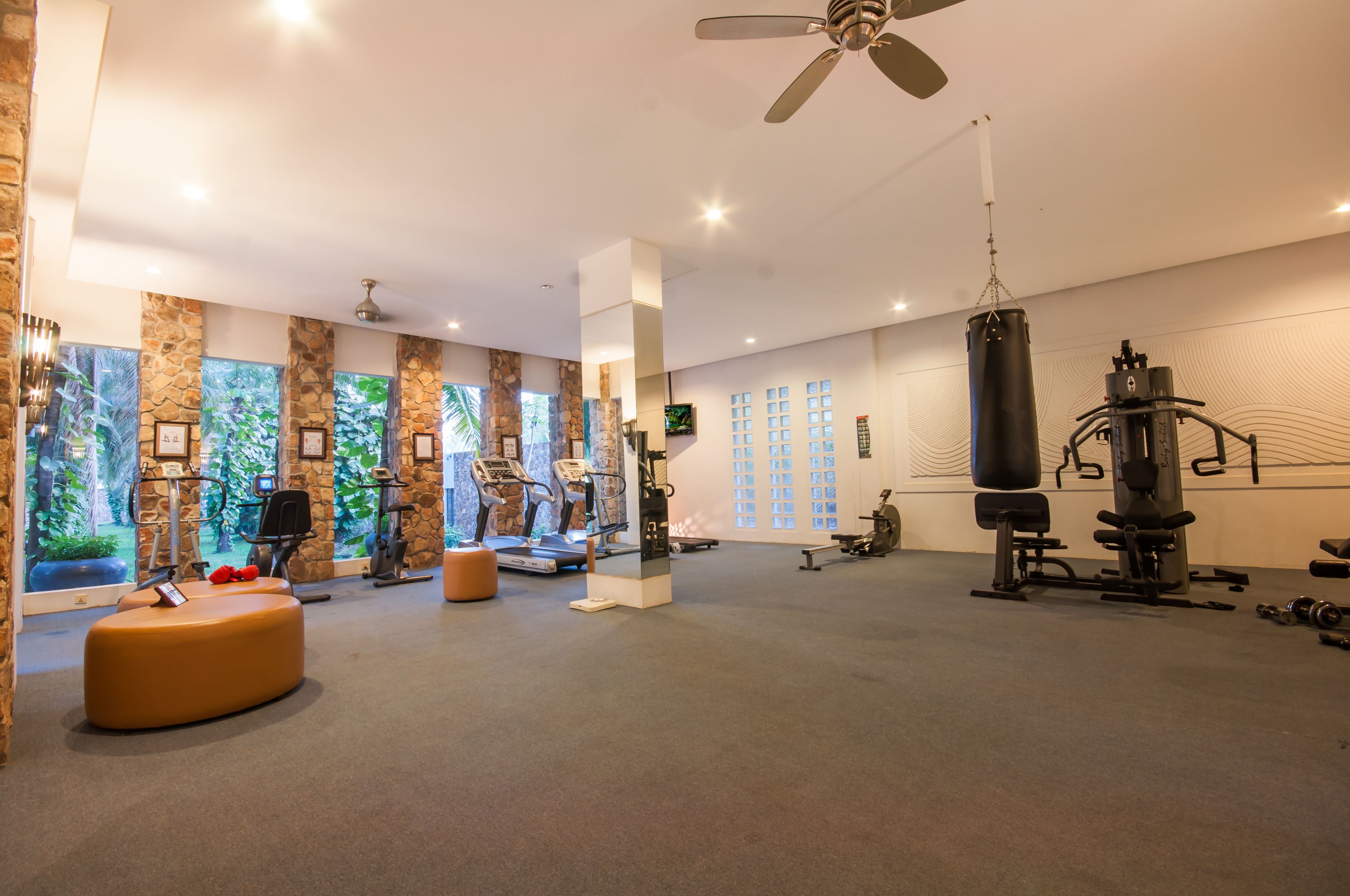 A spacious gym with various exercise equipment, a ceiling fan, and large windows with a view of greenery.