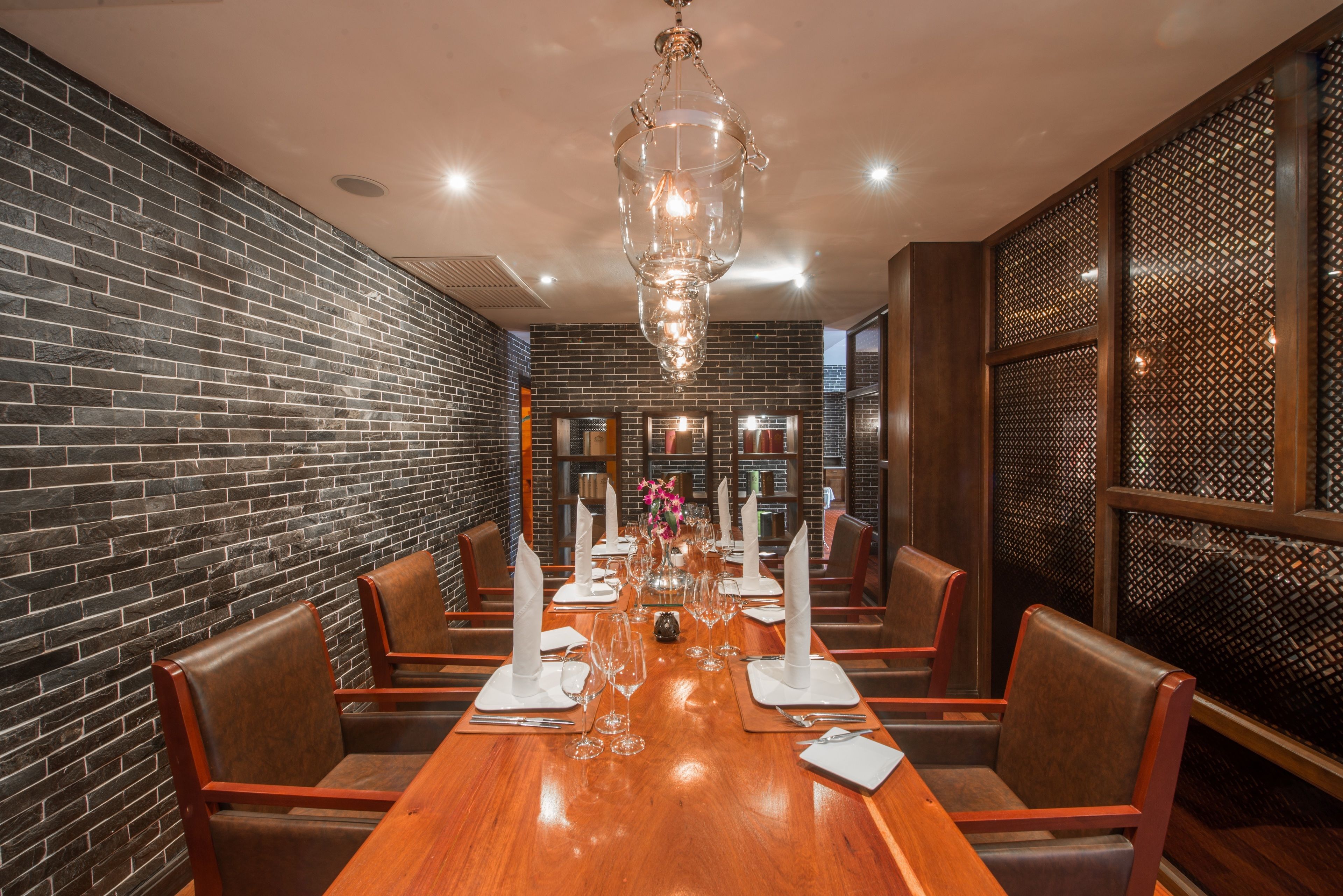 A dining area with a long wooden table set for six, surrounded by leather chairs, a chandelier, and brick walls.