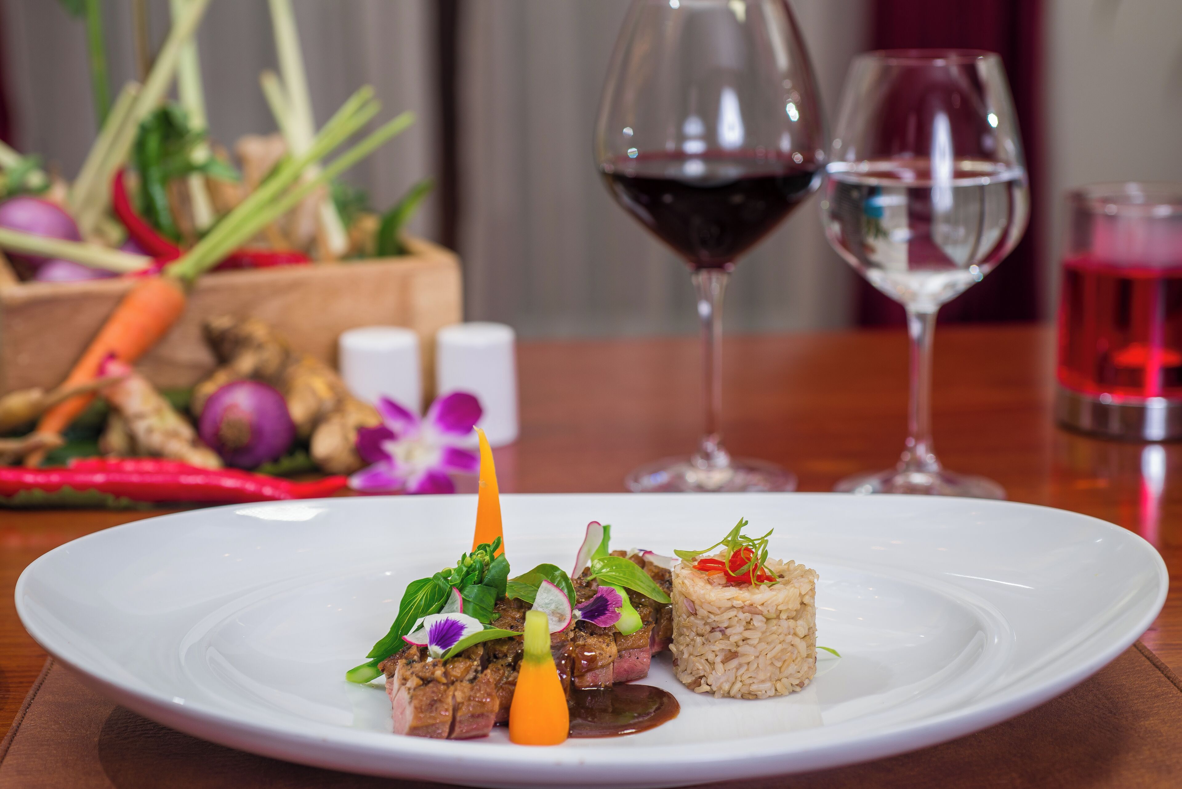 A white plate with a dish of meat, vegetables, and rice. There are two wine glasses in the background.