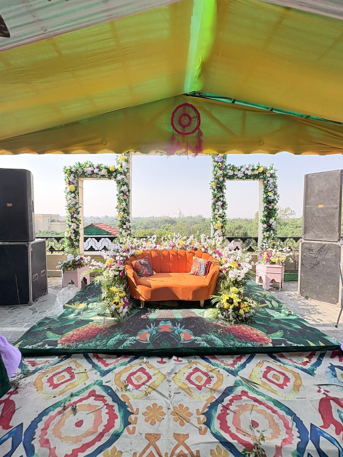 An outdoor seating area with a sofa under a canopy, surrounded by floral arrangements and a patterned floor.