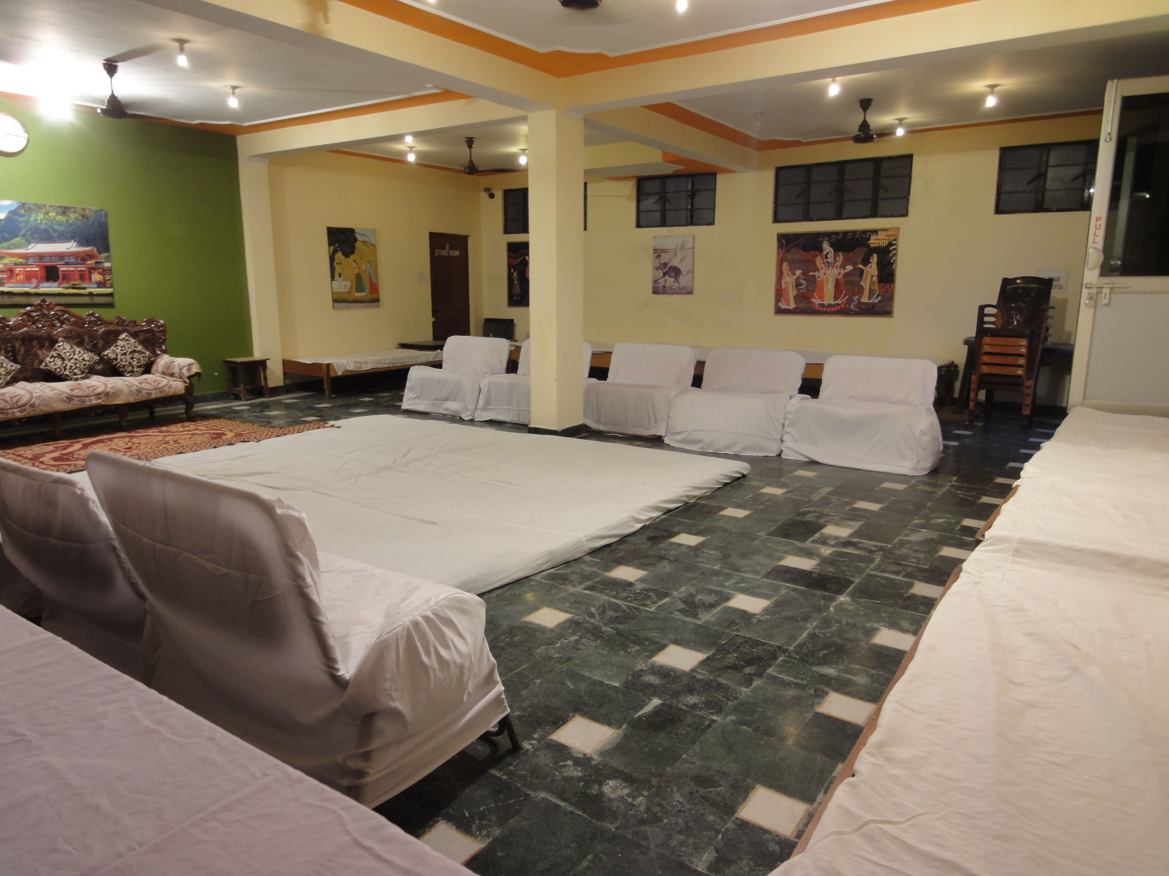 A hotel room with a large bed, a sofa, and several chairs. There are paintings on the walls and a ceiling fan.