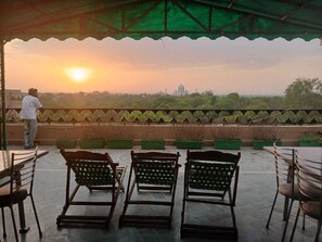 Une terrasse avec des chaises et une personne debout, d’où l’on peut admirer un paysage avec un bâtiment imposant à l’horizon.