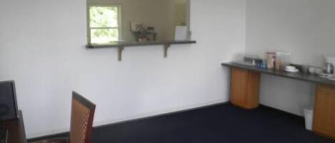 A room with a dark carpet, a wooden chair, a small table, a mirror, and a kitchenette area with cabinets and a countertop.